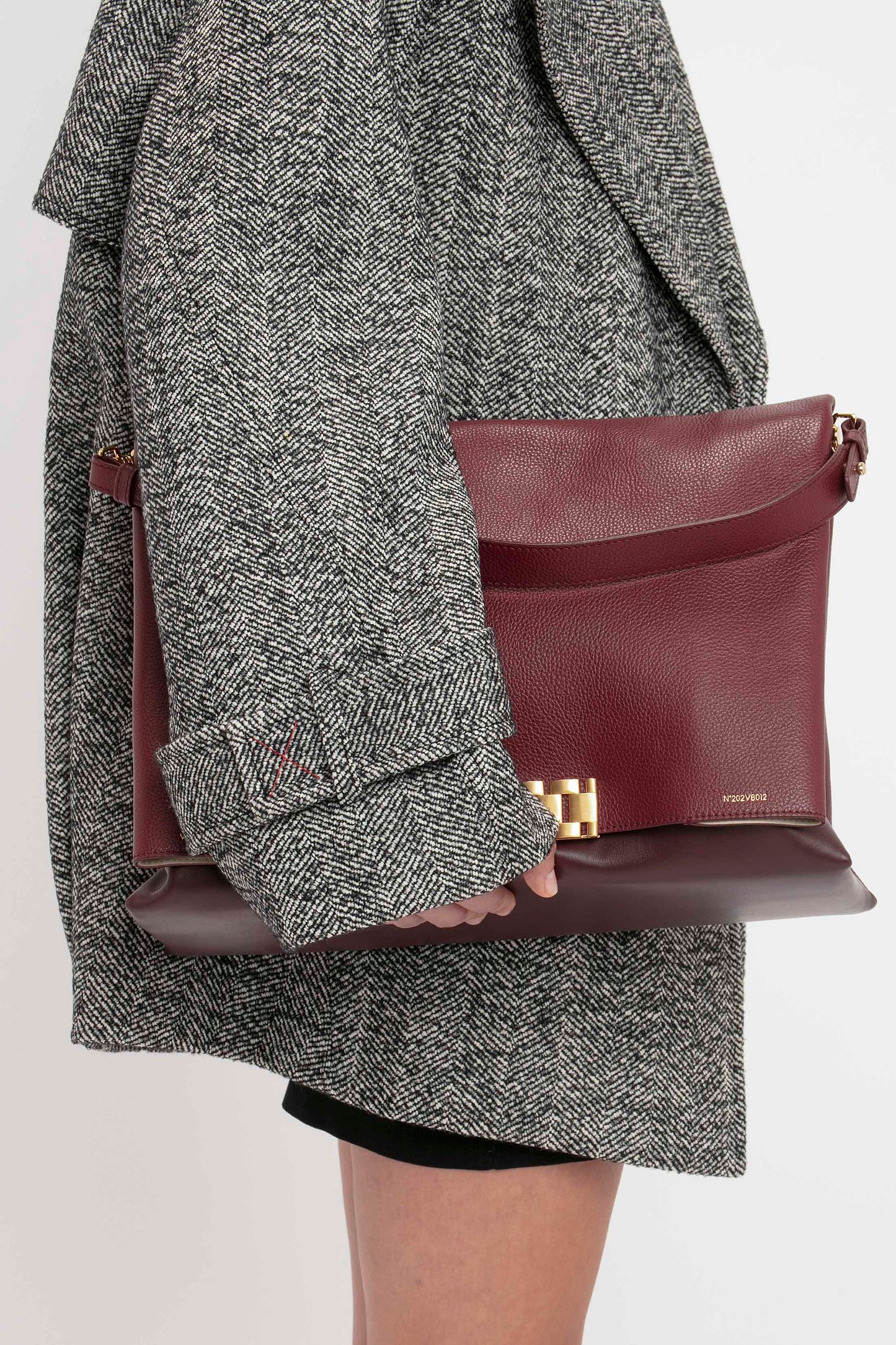 Person in a black VB Body Mini Dress by Victoria Beckham, holding a large maroon leather clutch bag with gold hardware against a white background.