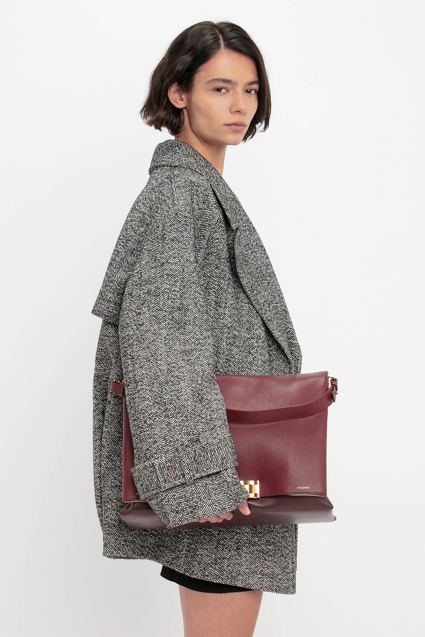 A person with short, dark hair wears the VB Body Mini Dress in Black by Victoria Beckham and holds a maroon clutch bag against a plain white background.