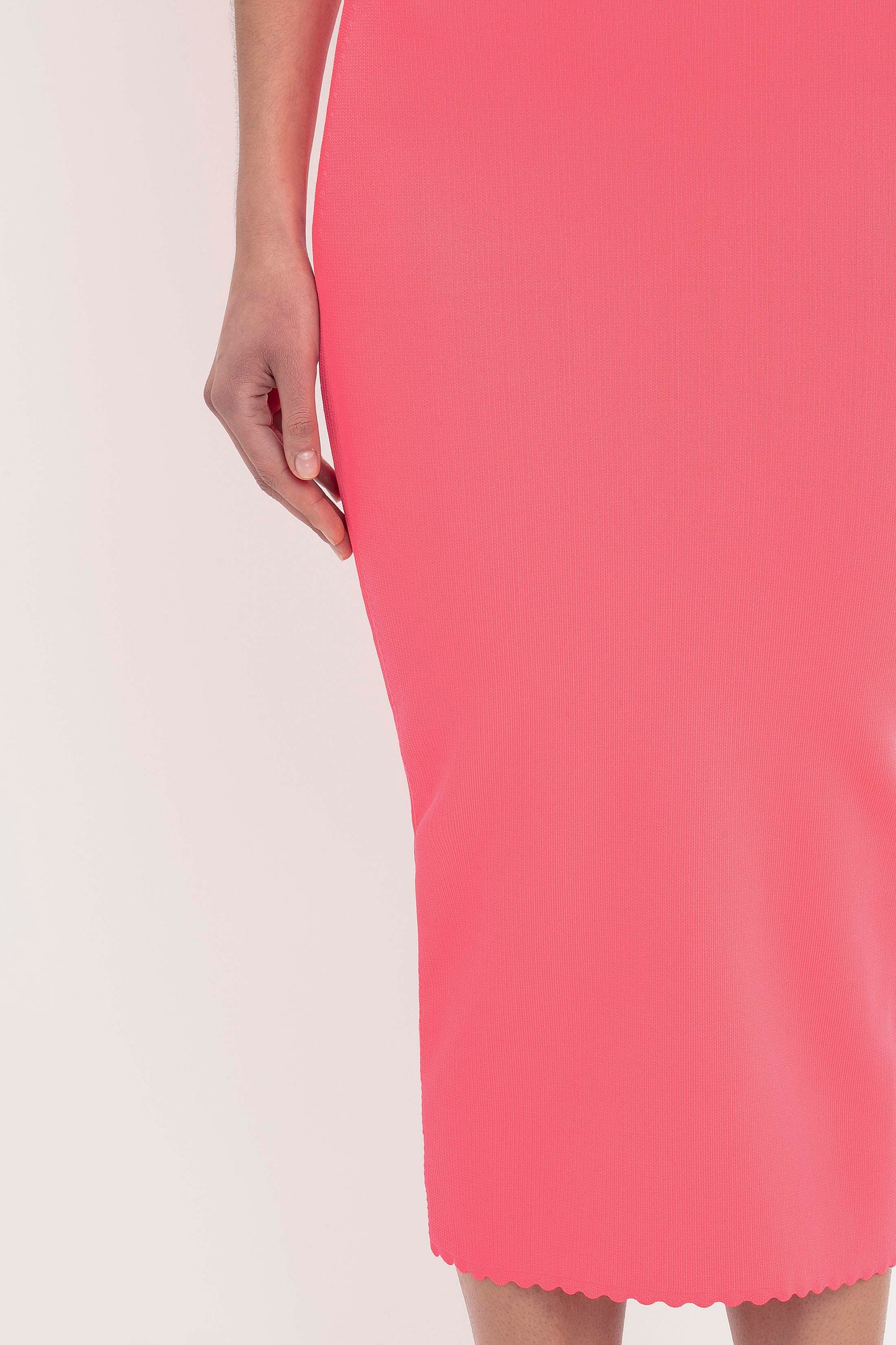Close-up of a person wearing the VB Body Slash Front Midi Dress in Pink by Victoria Beckham. The fitted, knee-length dress features a scalloped hem, with only the lower torso, hand, and part of the legs visible against a plain white background.