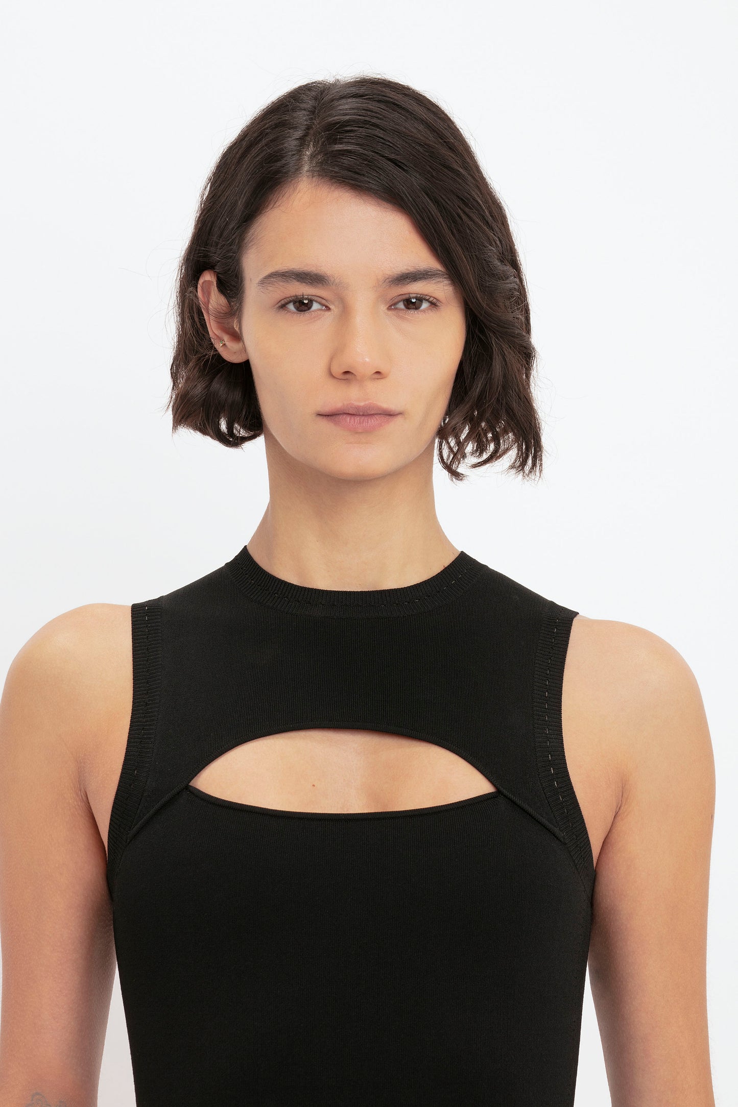 A person with short, dark hair wearing a sleeveless VB Body Cut Out Midi Dress in Black by Victoria Beckham.