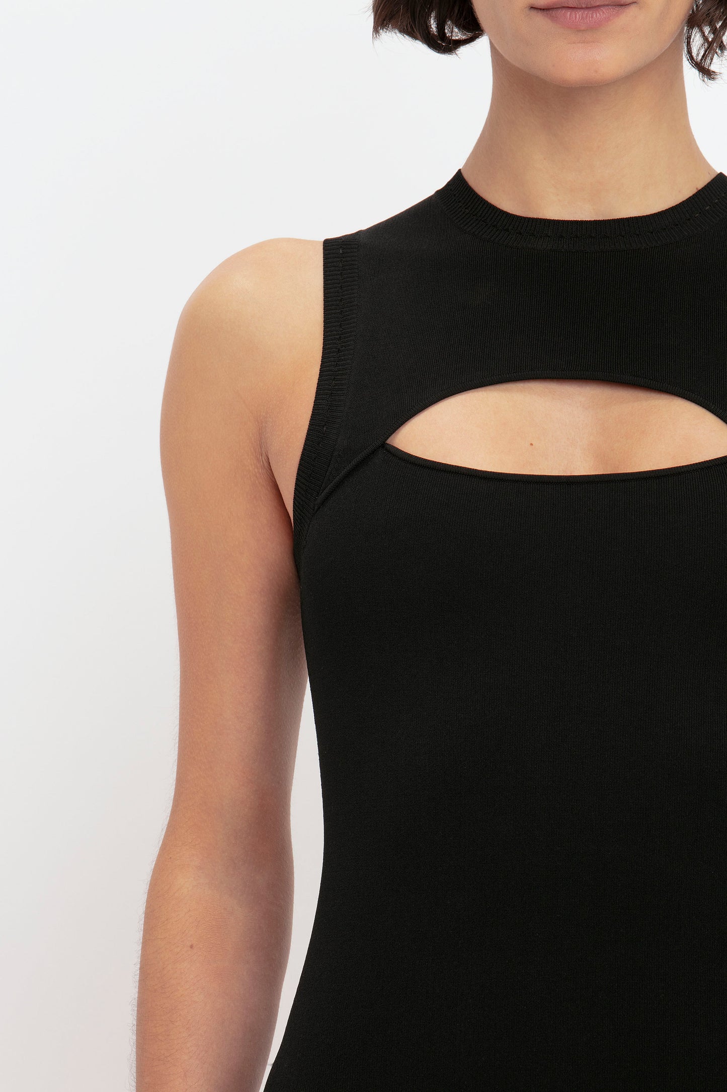 A person wearing the VB Body Cut Out Midi Dress in Black by Victoria Beckham, showcasing a sleek sleeveless design with a chic cut-out detail on the chest area, visible from the shoulders to just above the waist.