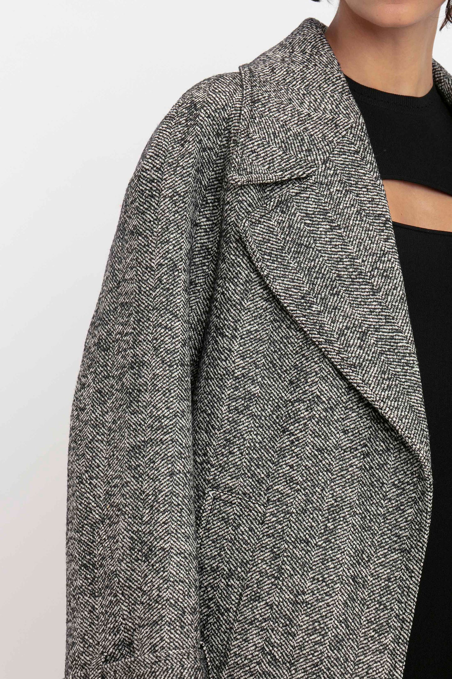 Close-up of a person wearing a grey herringbone tweed coat with wide lapels, over the Victoria Beckham VB Body Cut Out Midi Dress in Black with a horizontal cutout detail on the chest.