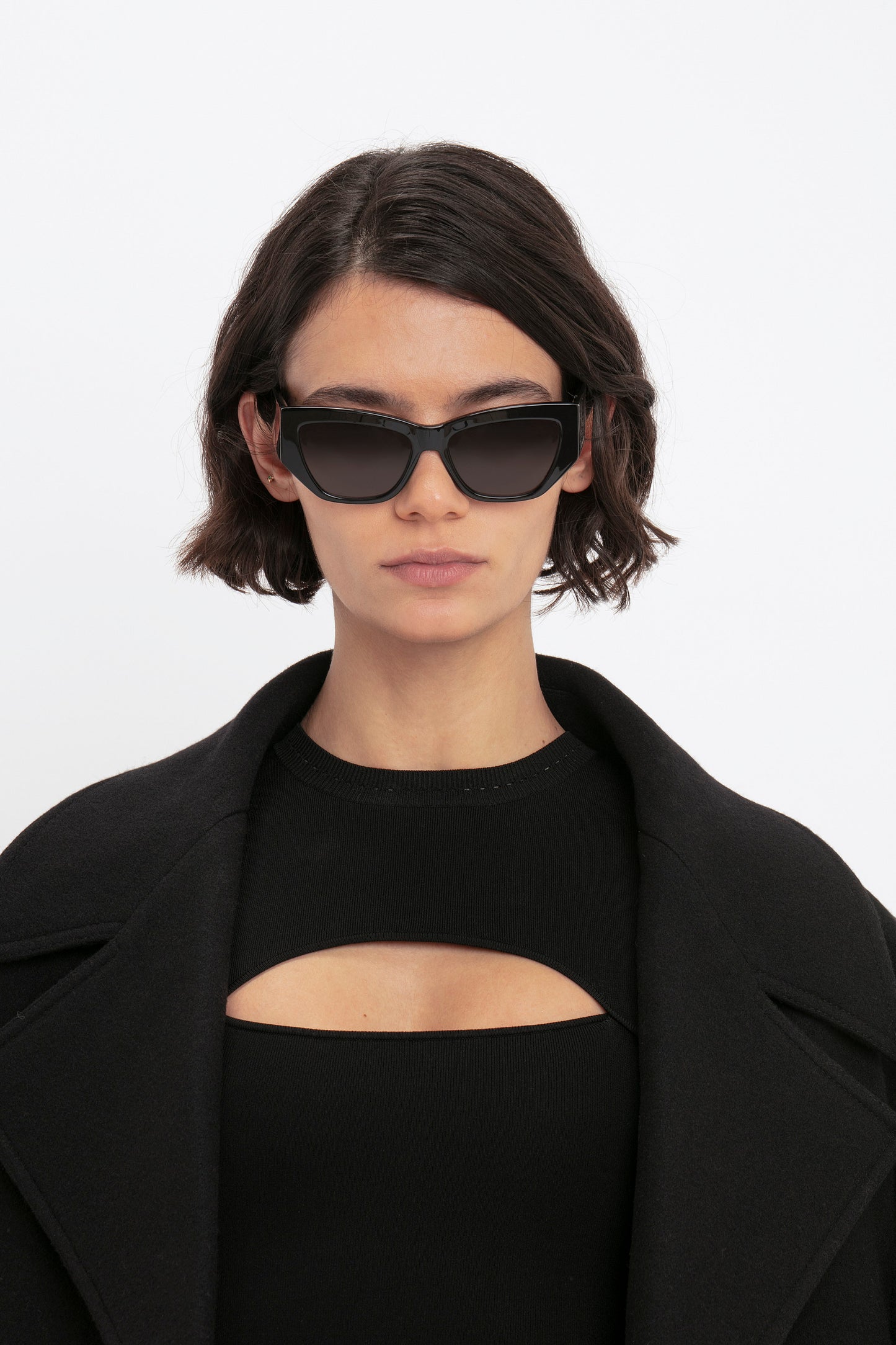 Person wearing dark sunglasses, a black coat, and the VB Body Cut Out Midi Dress in Black from Victoria Beckham, standing against a plain white background.
