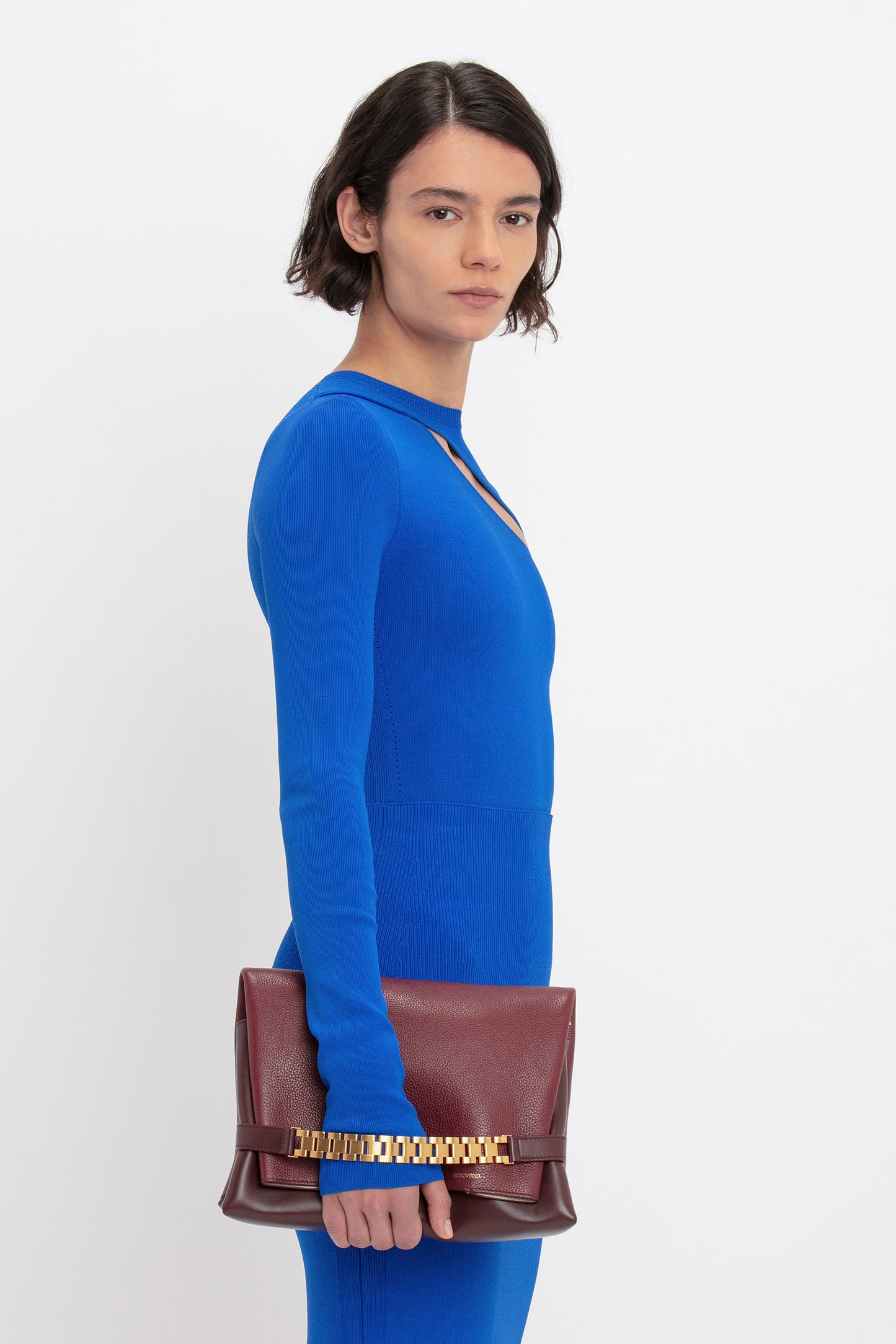 A person wearing the VB Body Front Slash Crew Neck Top in Cobalt from Victoria Beckham holds a brown clutch with a gold chain detail against a plain background.