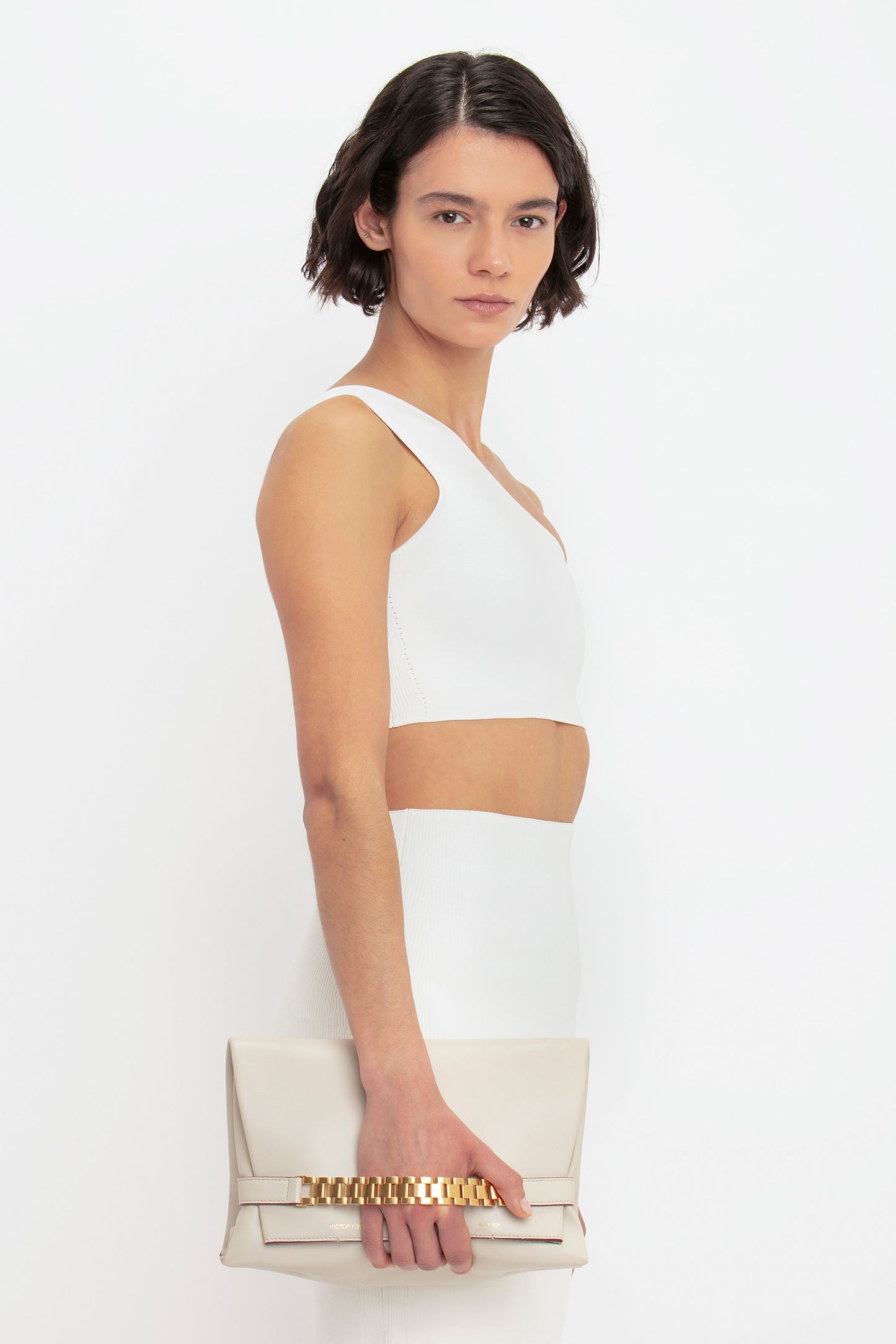 A person with short dark hair is wearing the Victoria Beckham VB Body One Shoulder Crop Top in White and a matching skirt, holding a beige clutch with a gold chain detail. They are posing against a plain white background.