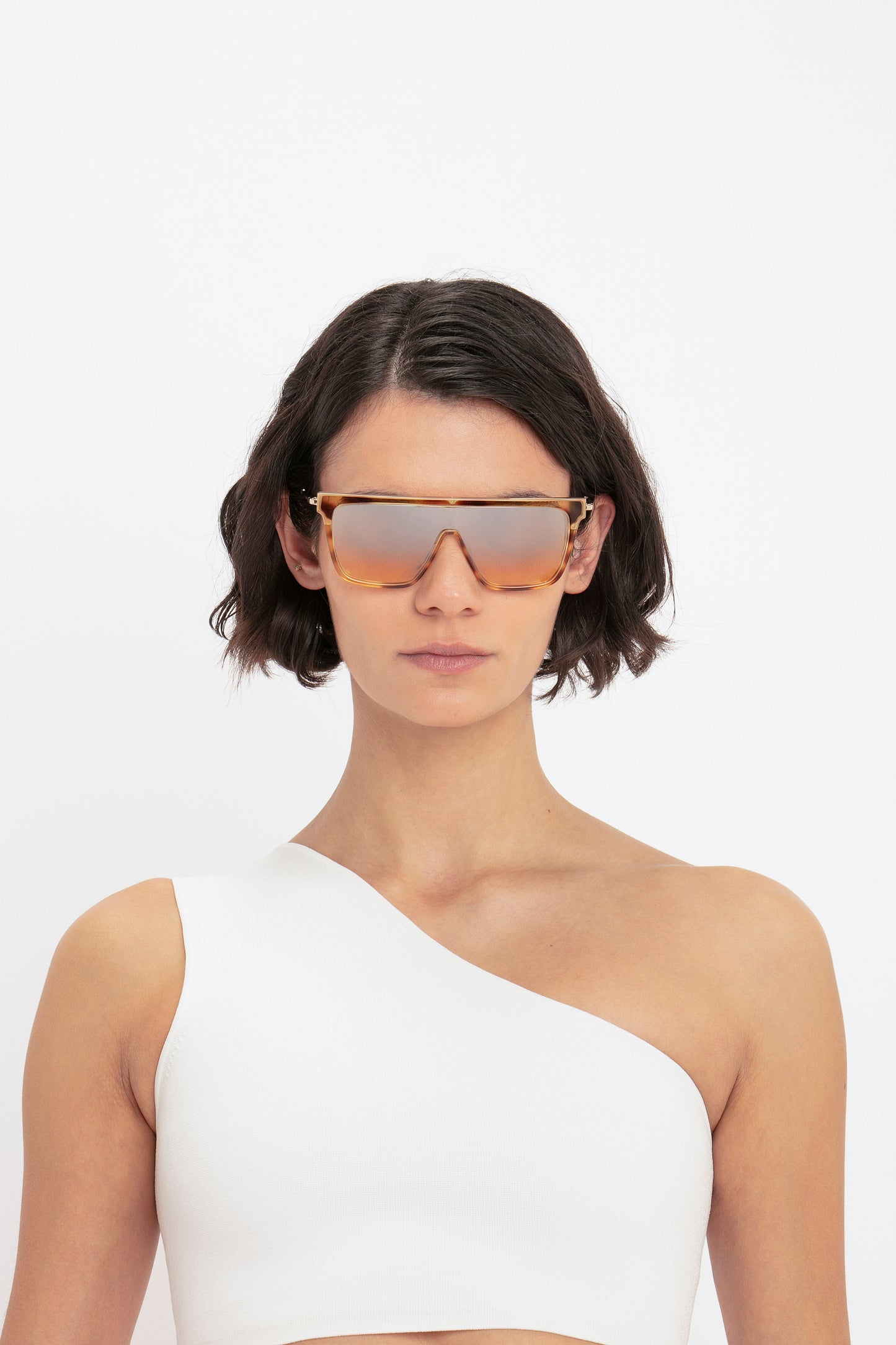 A person with short dark hair wearing large, reflective sunglasses and the VB Body One Shoulder Crop Top in White by Victoria Beckham against a plain white background.