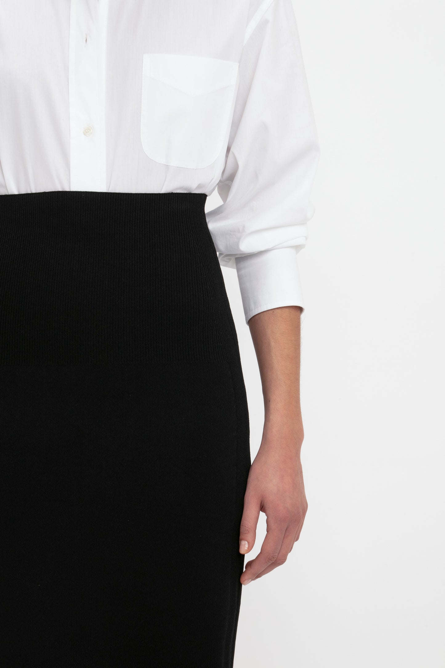 A person wears a white button-down shirt tucked into the high-waisted, black VB Body Fitted Midi Skirt by Victoria Beckham. Only the torso and right arm are visible.