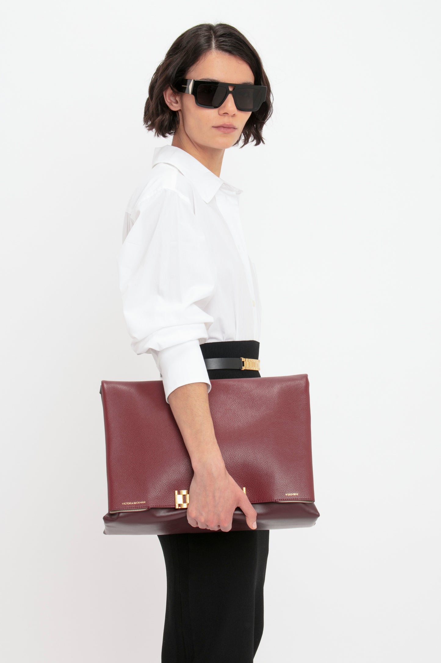 A person dressed in a white shirt, Victoria Beckham's VB Body Fitted Midi Skirt in Black, and large sunglasses is holding a large maroon clutch bag against a plain white background.