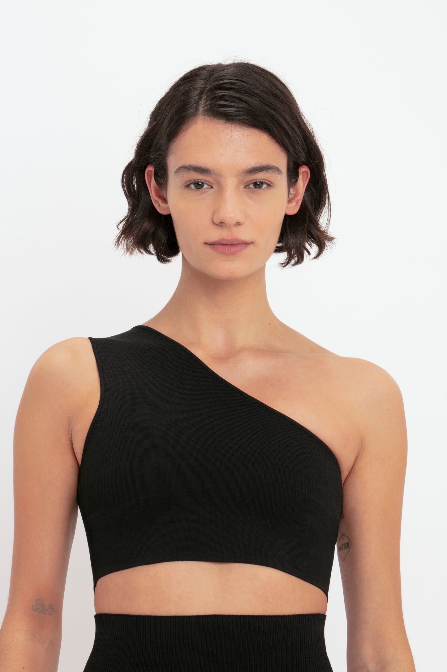 A woman with short dark hair wearing the VB Body One Shoulder Crop Top in Black by Victoria Beckham stands against a plain white background.