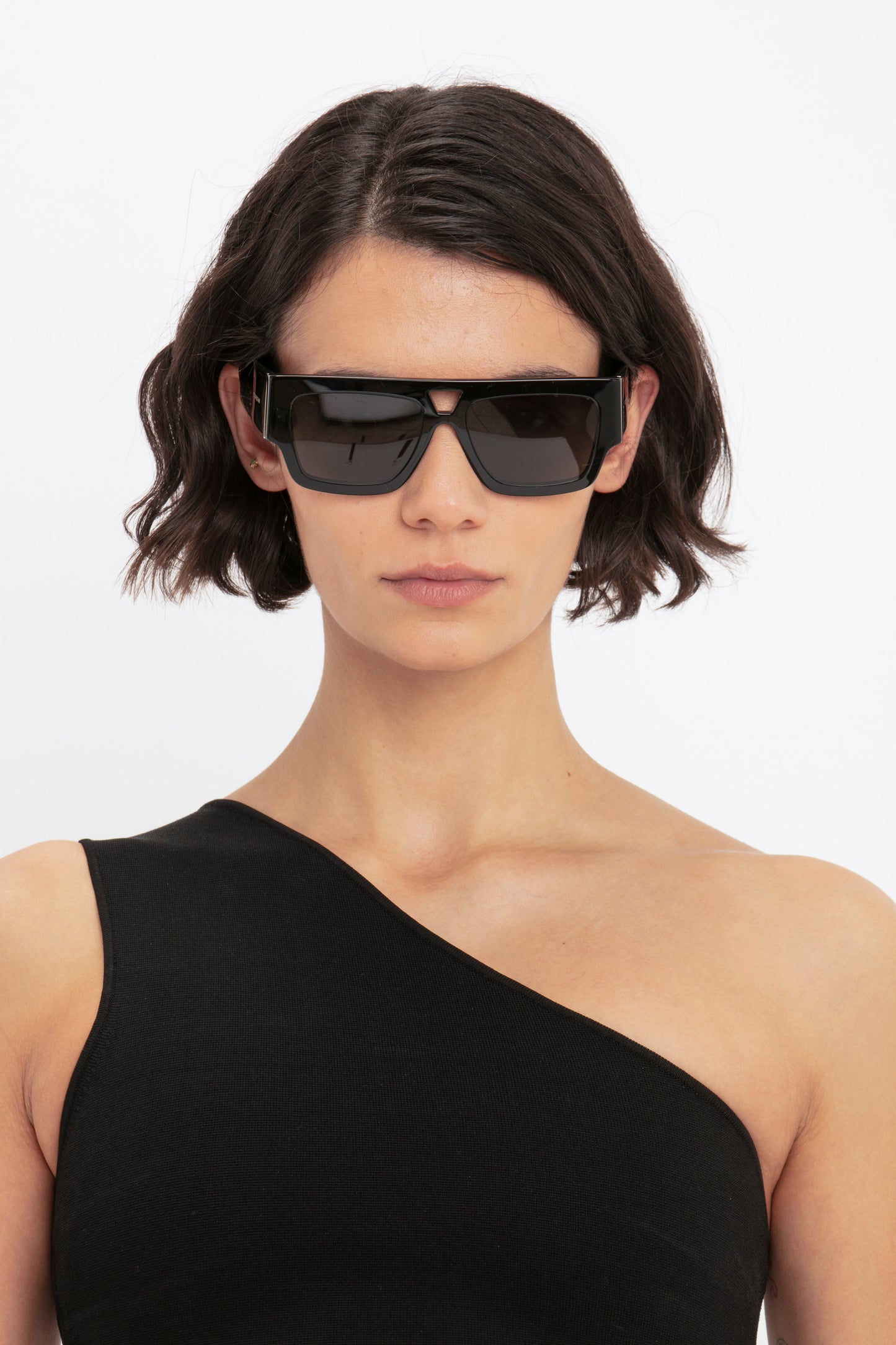 Person with short, dark hair wearing black sunglasses and the Victoria Beckham VB Body One Shoulder Crop Top in black, posing against a plain white background.