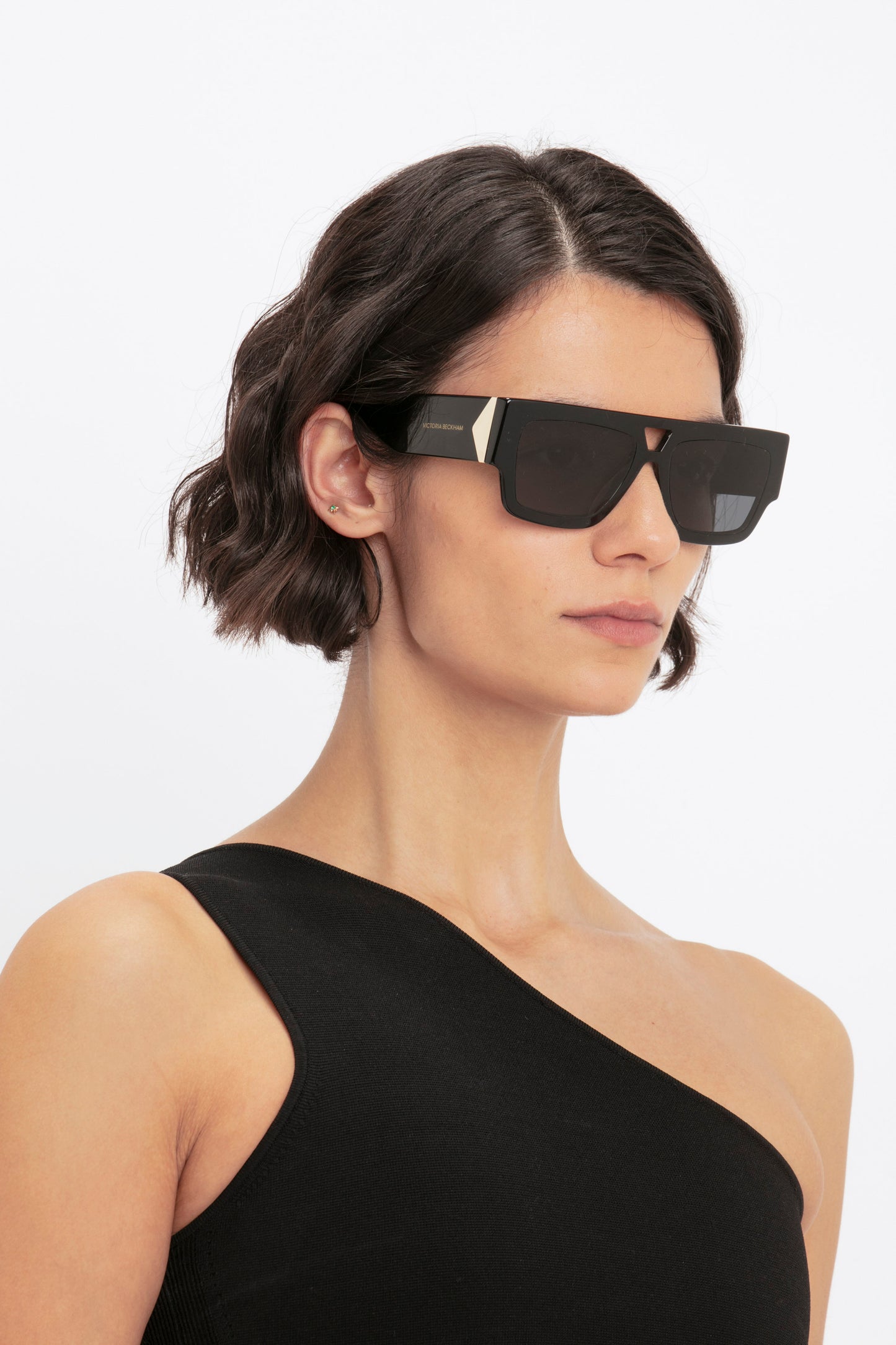 A person with short, wavy dark hair wearing large black sunglasses and the VB Body One Shoulder Crop Top in Black by Victoria Beckham, looking ahead.