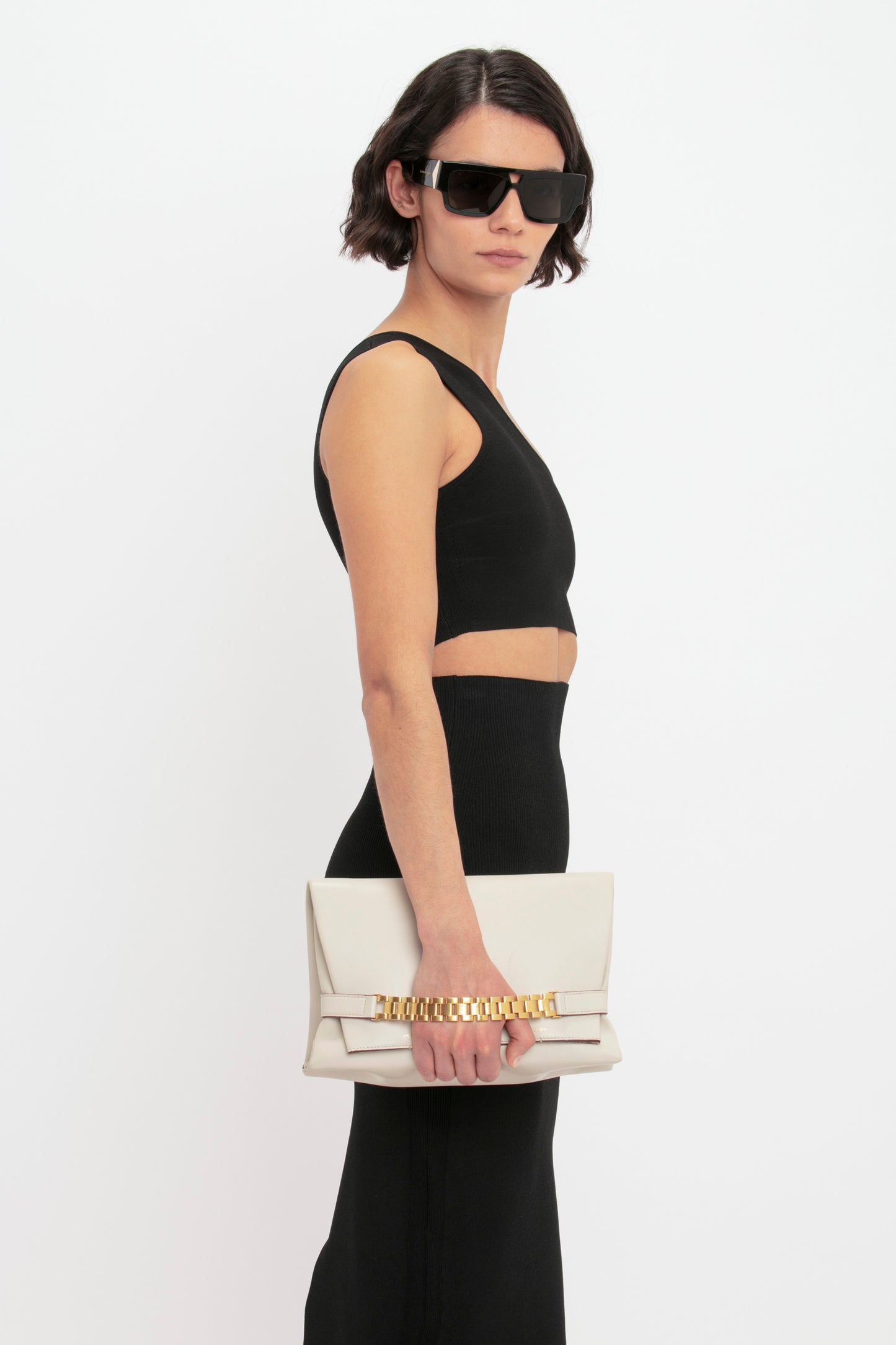 A woman wearing large black sunglasses, a black crop top, and a VB Body Fitted Midi Skirt in Black by Victoria Beckham holds a beige clutch purse.