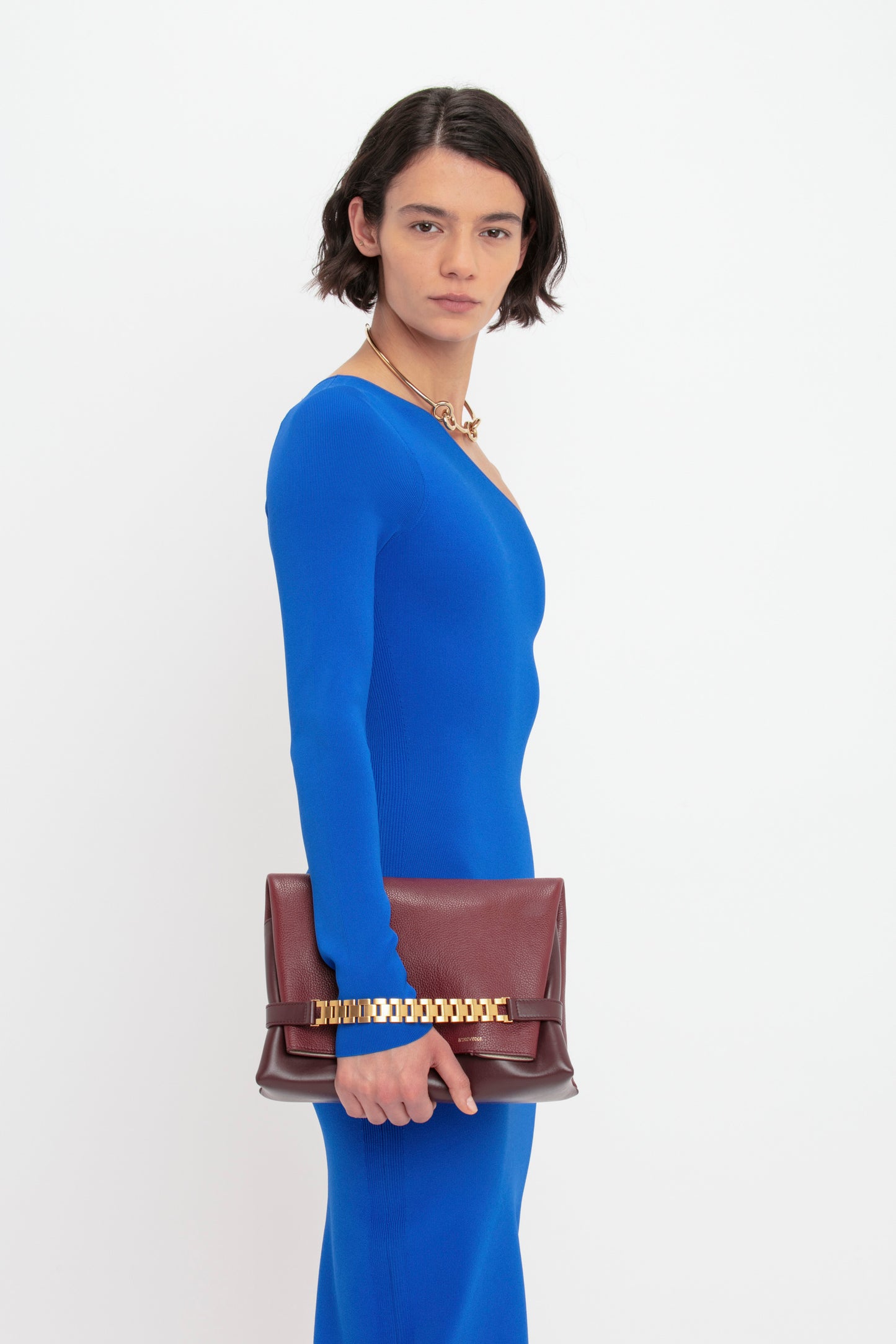 A person stands against a white background, wearing the Victoria Beckham VB Body One Shoulder Midi Dress in Cobalt and holding a brown clutch.