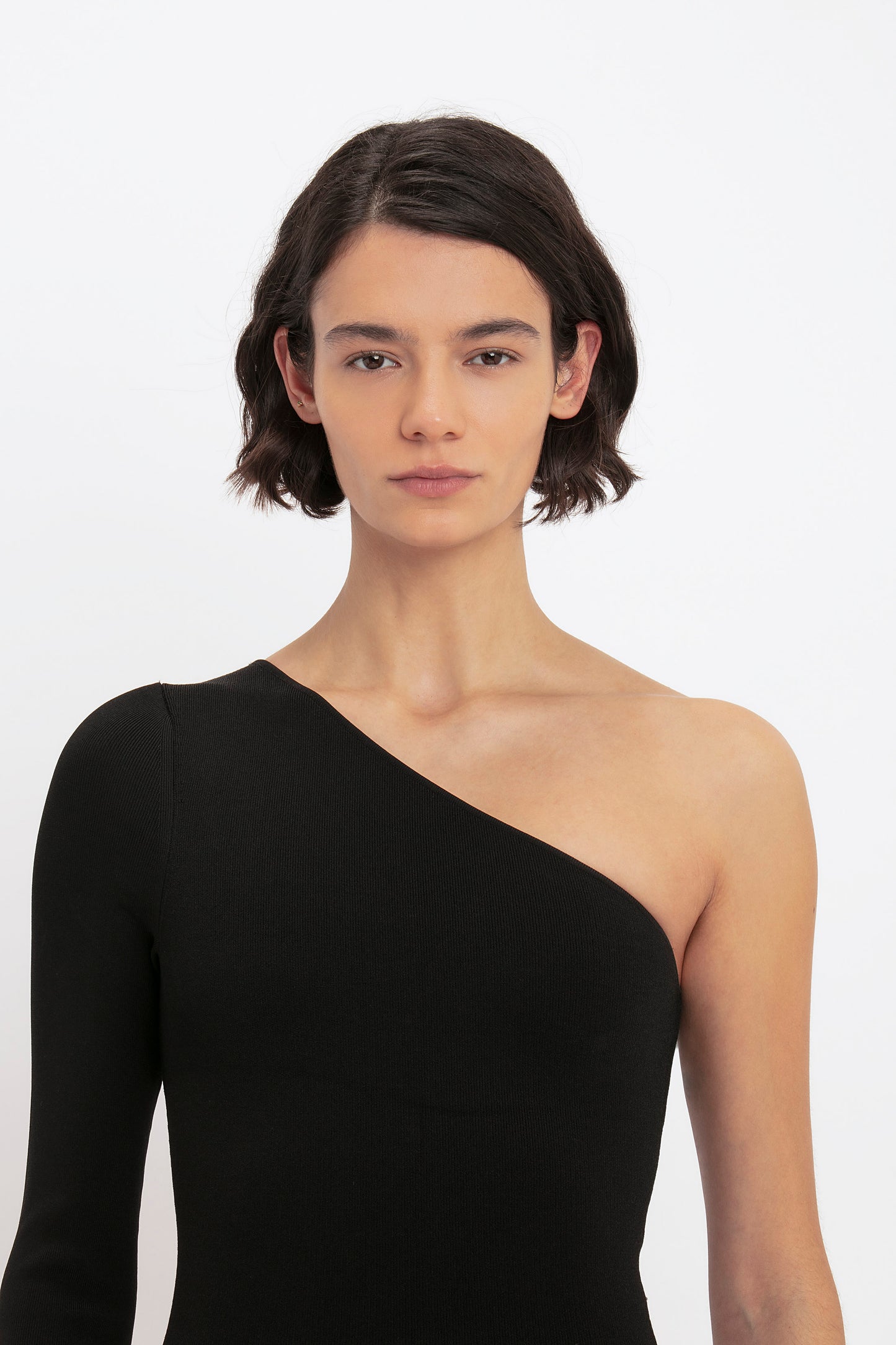 Person with short dark hair wearing the VB Body One Shoulder Midi Dress in Black by Victoria Beckham, looking straight ahead against a plain background.