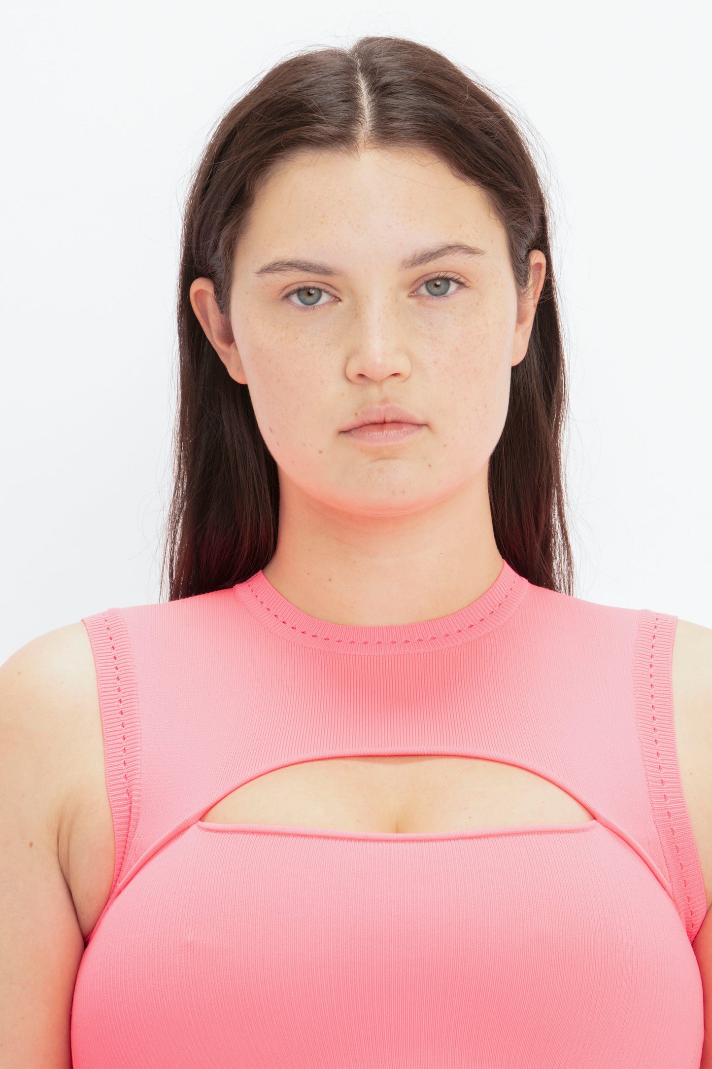 A woman with long dark hair wears the VB Body Slash Front Midi Dress in Pink by Victoria Beckham, featuring a cutout detail on the chest. She stands facing the camera against a plain, white background.