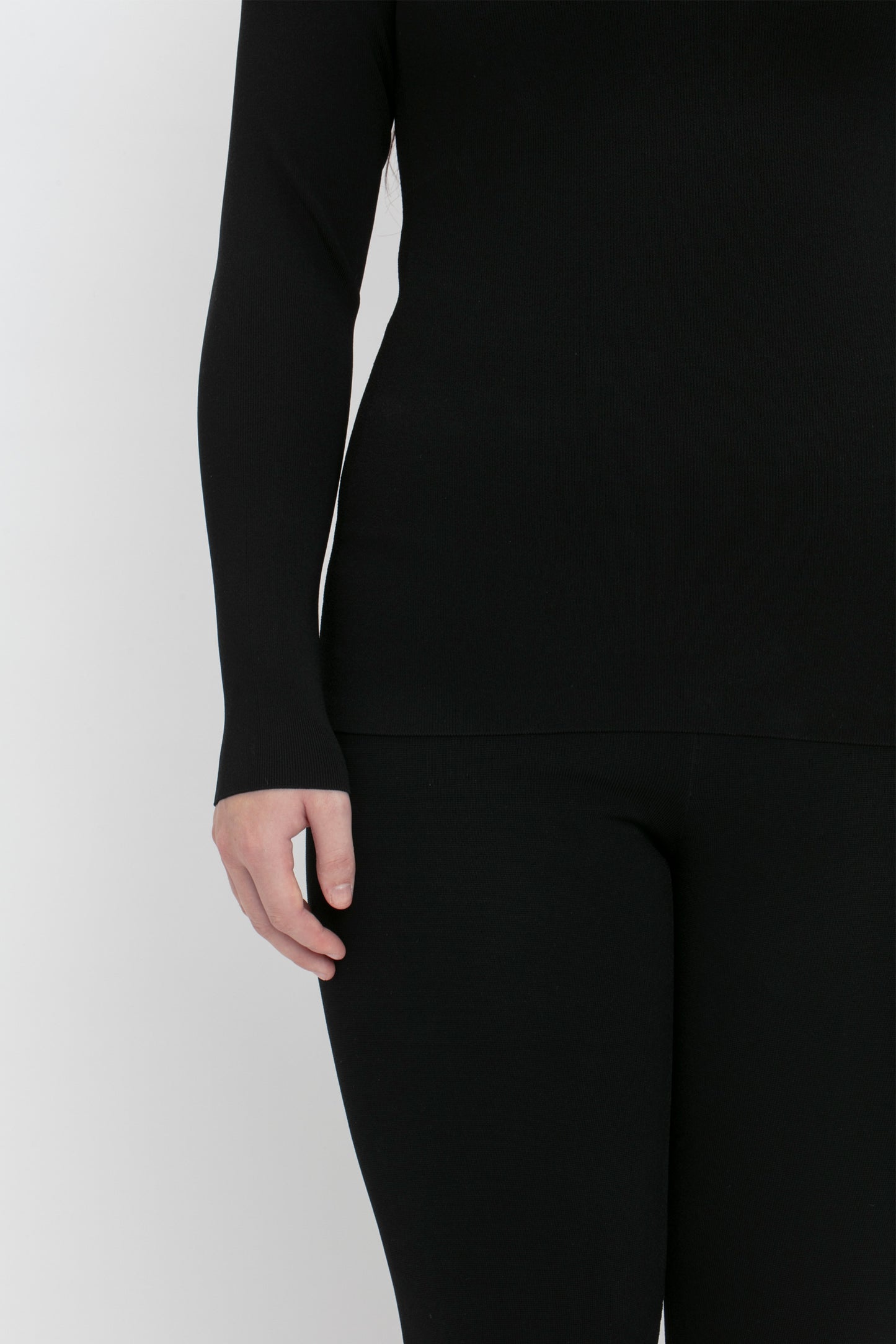 A person dressed in a black VB Body One Shoulder Top by Victoria Beckham and black pants, with their right hand partially visible at their side. The background is plain white.