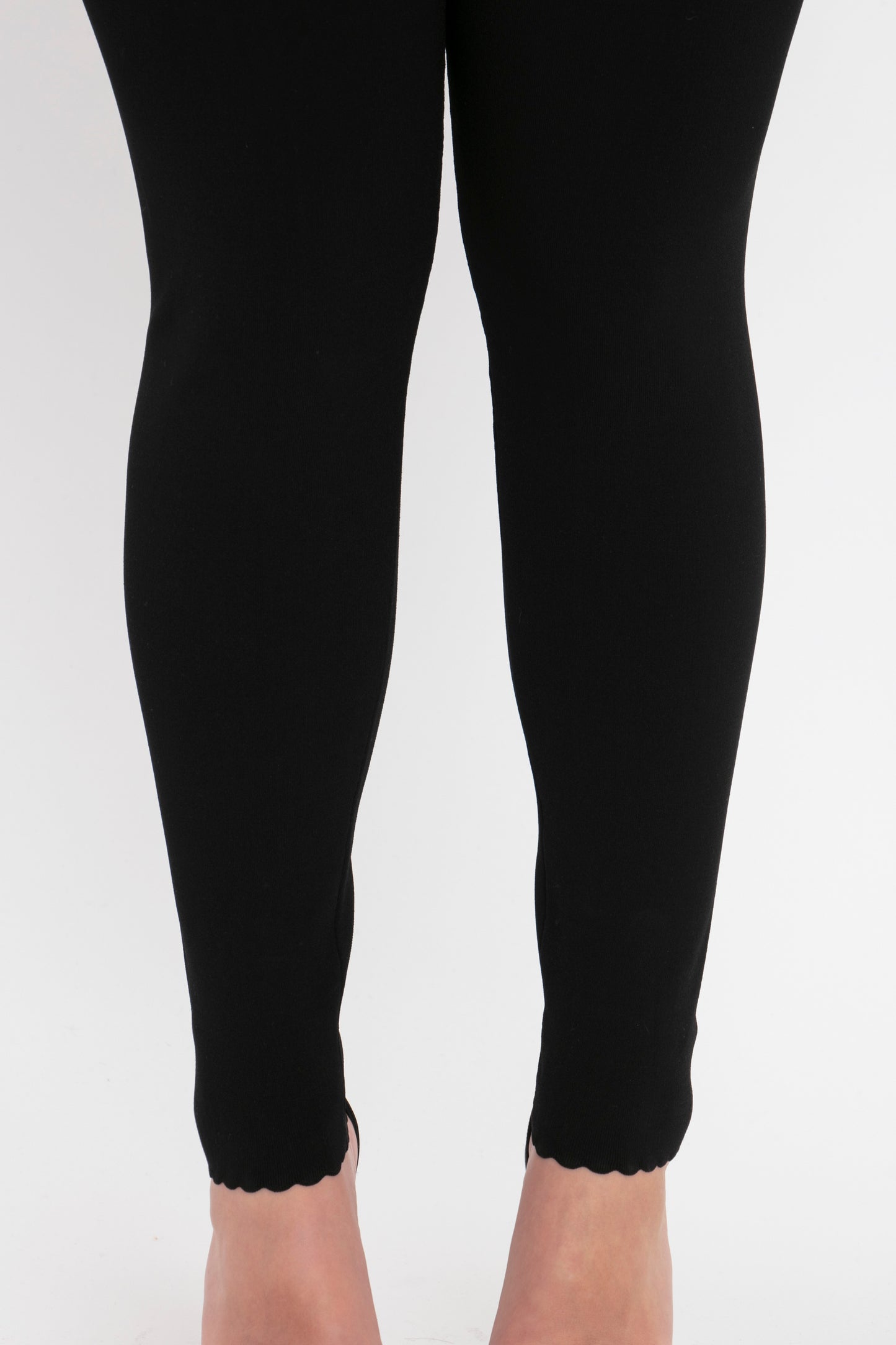 Close-up of a person's lower legs wearing the VB Body Catsuit in Black by Victoria Beckham. The catsuit ends just above the ankles, exposing the feet. The background is white.