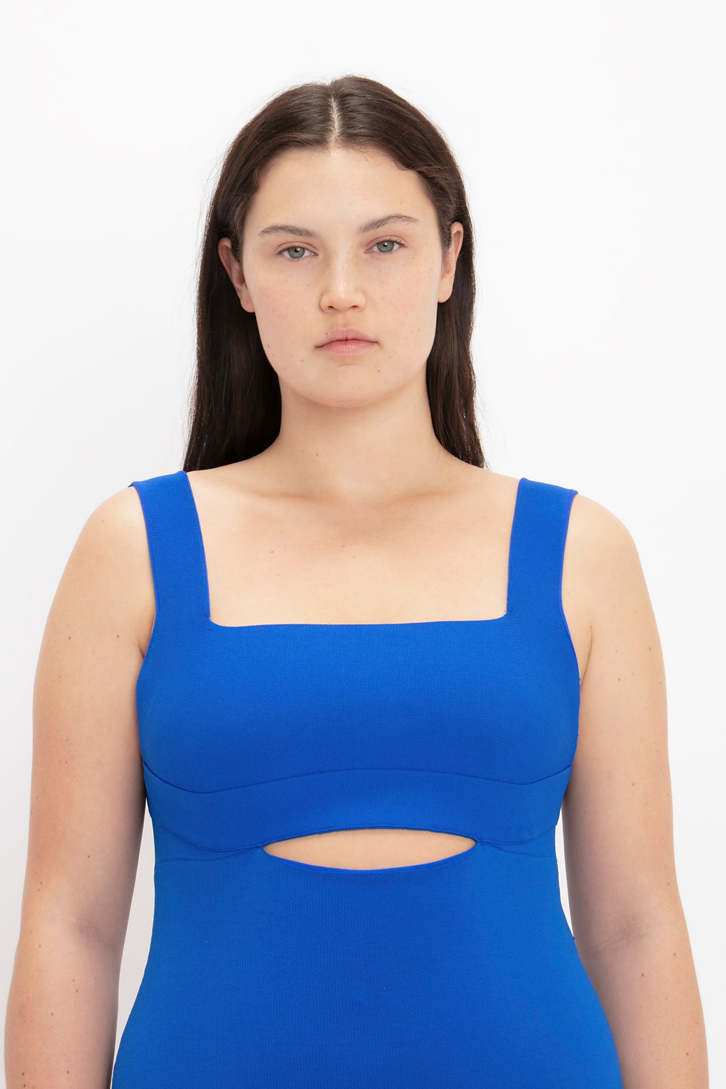A woman with straight dark hair and a neutral expression is wearing the VB Body Cut Out Midi Dress in Cobalt by Victoria Beckham, which features a bright blue hue, sleeveless design, square neckline, and a cutout at the front.
