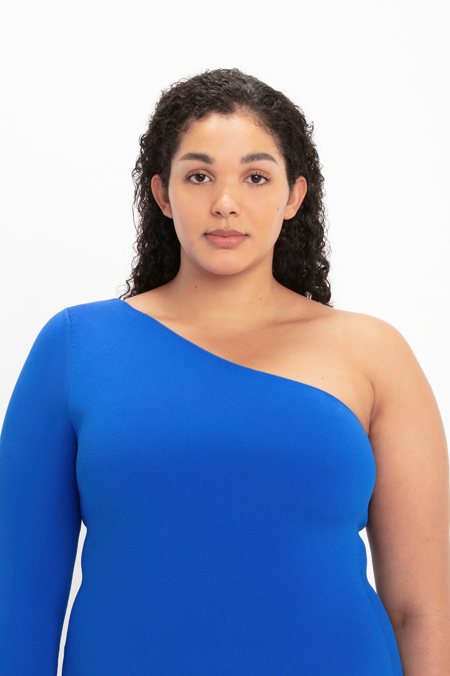 A person with curly hair wearing the Victoria Beckham VB Body One Shoulder Midi Dress in Cobalt stands against a plain white background.