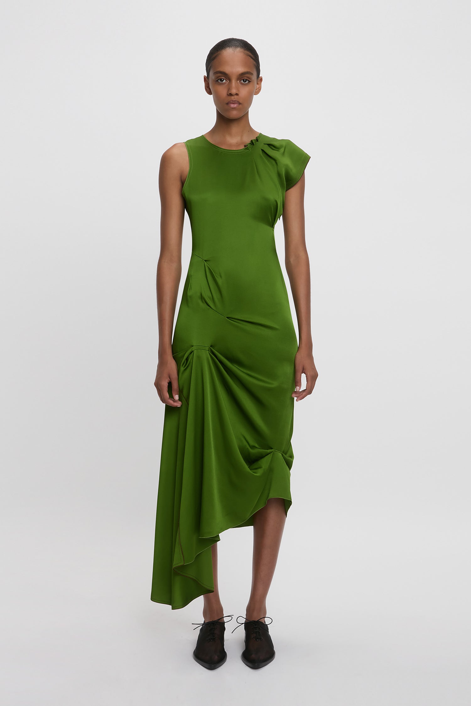 Someone wears the Victoria Beckham Draped Tuck Detail Midi Dress in Algae Green, featuring asymmetrical ruffles like the Spring Summer 2025 runway style, paired with black shoes against a plain white background.