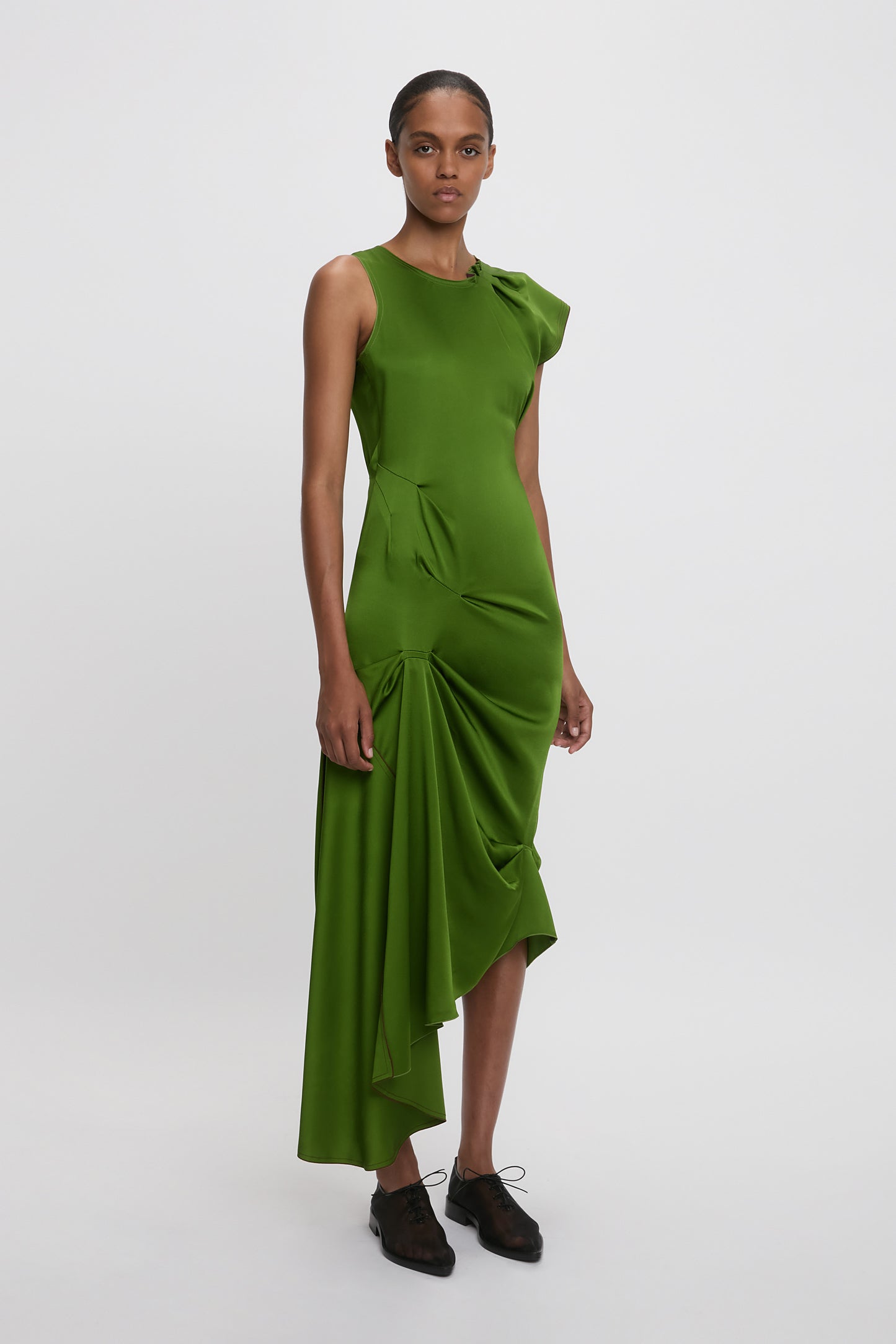The person is wearing a Victoria Beckham Draped Tuck Detail Midi Dress in algae green with black shoes, standing against a plain white background.
