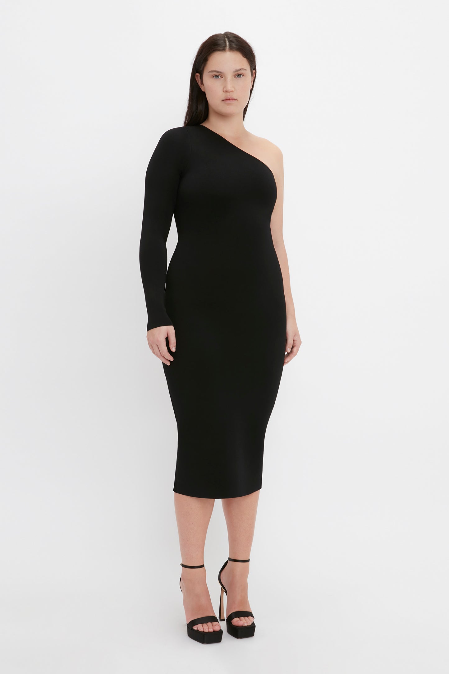 A woman stands against a plain white background, wearing the "VB Body One Shoulder Midi Dress in Black" by Victoria Beckham. She pairs the black bodycon dress with high heels and has straight, dark hair.