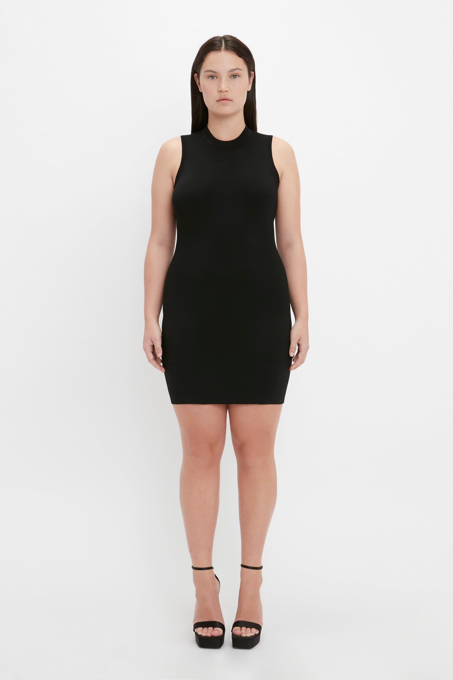 A person standing against a white background, wearing the Victoria Beckham VB Body Mini Dress in Black and black high-heeled shoes.