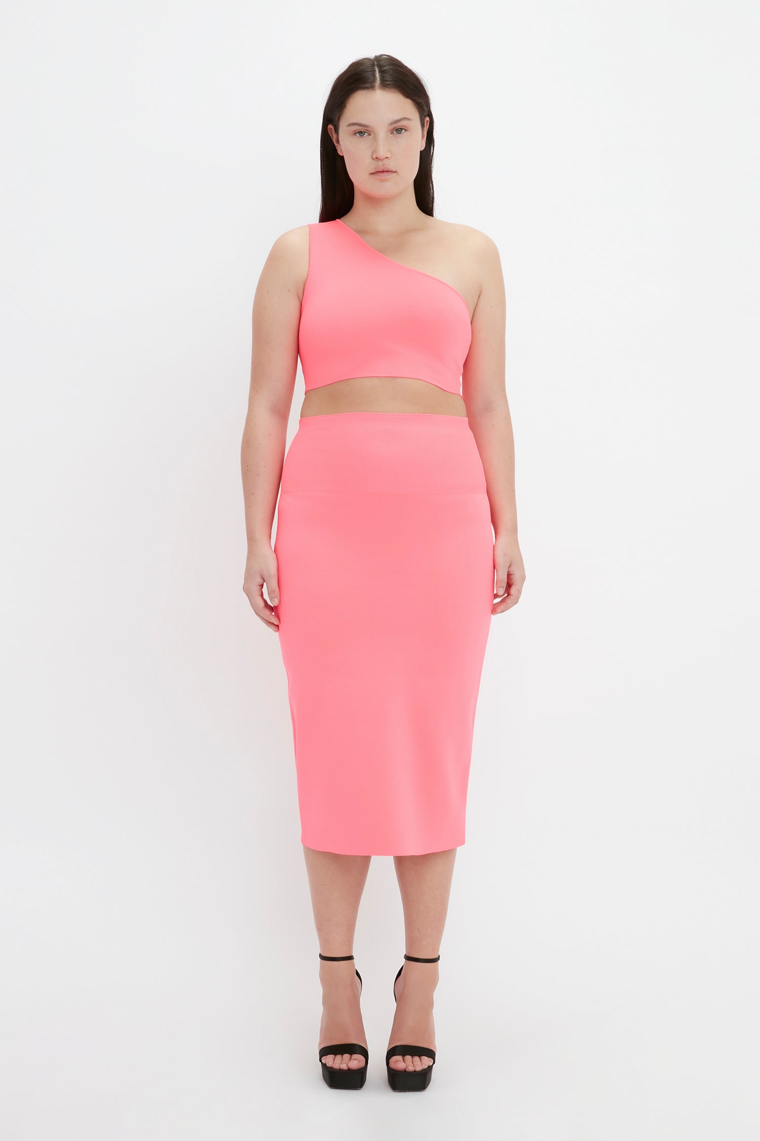 A person stands against a plain background wearing the VB Body One Shoulder Crop Top in Pink by Victoria Beckham and a matching high-waisted skirt, paired with black high heels.
