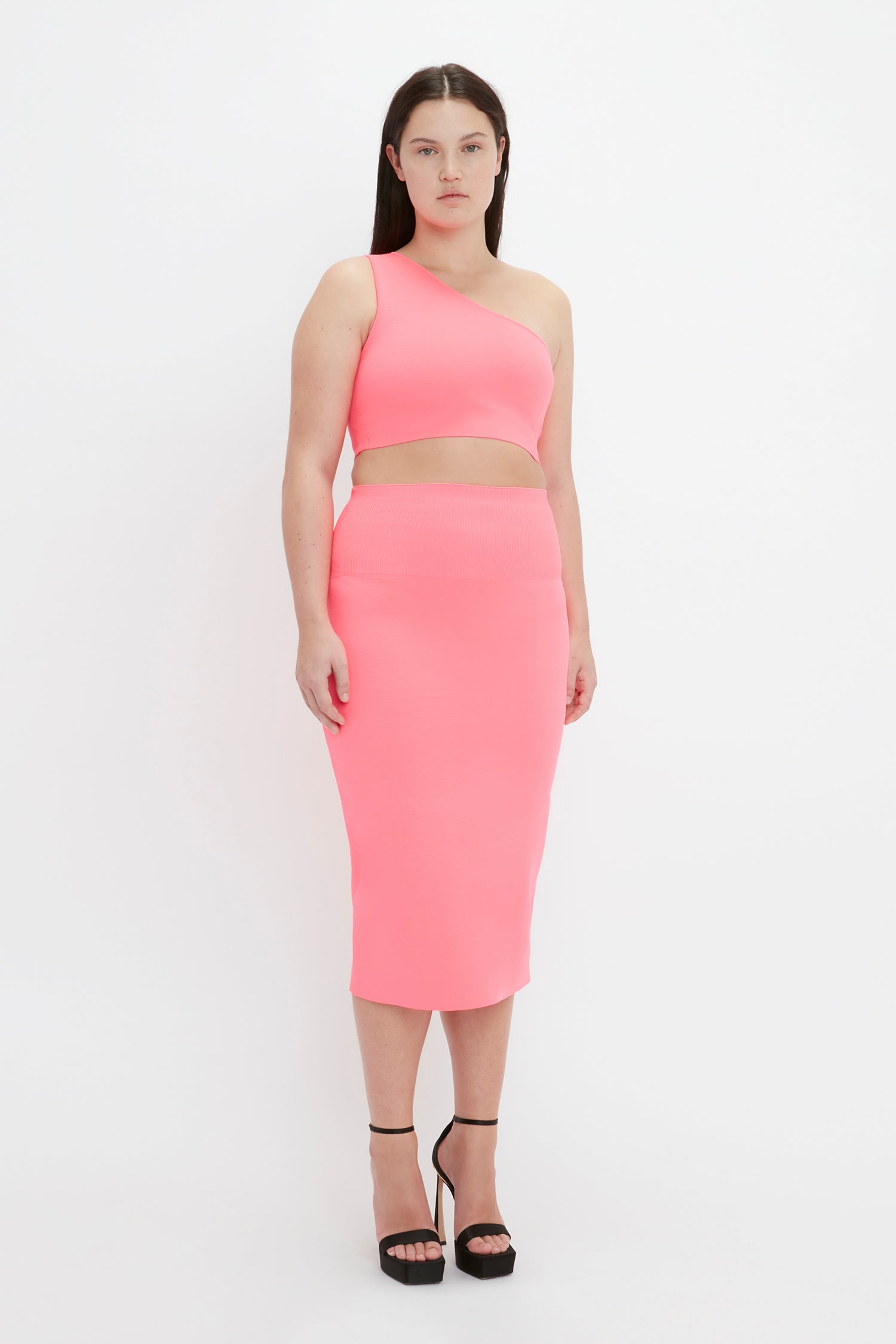 A woman stands facing forward, wearing the Victoria Beckham VB Body One Shoulder Crop Top in Pink, paired with a matching high-waisted skirt and black high heels.