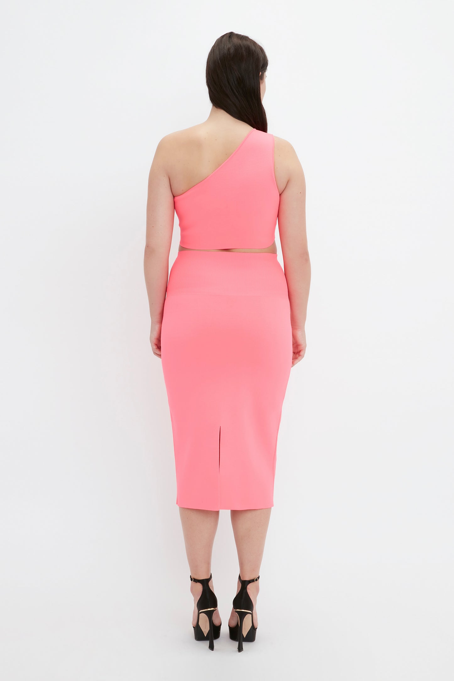 A woman is standing facing away, wearing the VB Body One Shoulder Crop Top in Pink by Victoria Beckham along with a matching pink skirt featuring a slit. She completes her outfit with black high-heeled sandals.