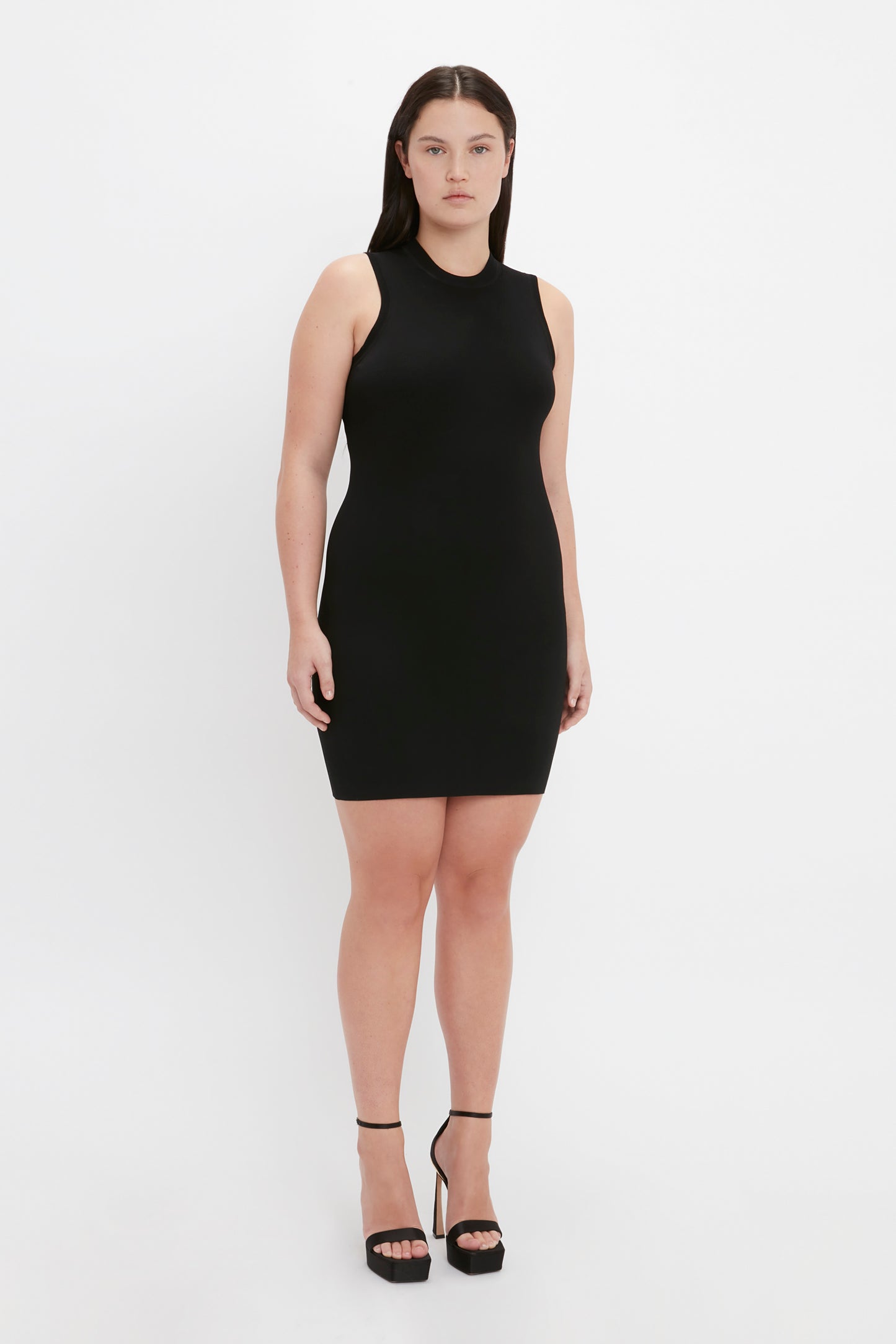 A person is standing against a plain white background wearing the sleeveless, black, form-fitting VB Body Mini Dress by Victoria Beckham and black high-heeled shoes.