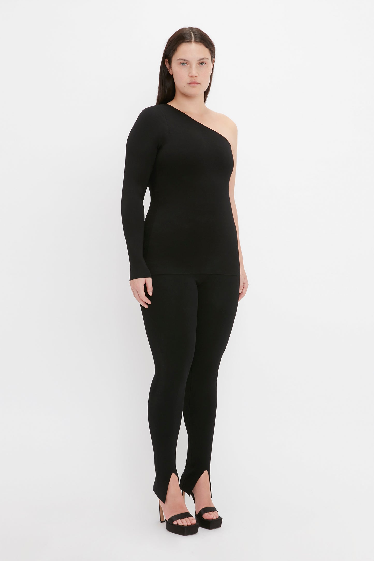 A woman stands against a white background, wearing the VB Body One Shoulder Top in black by Victoria Beckham, paired with long pants and black high-heeled shoes.