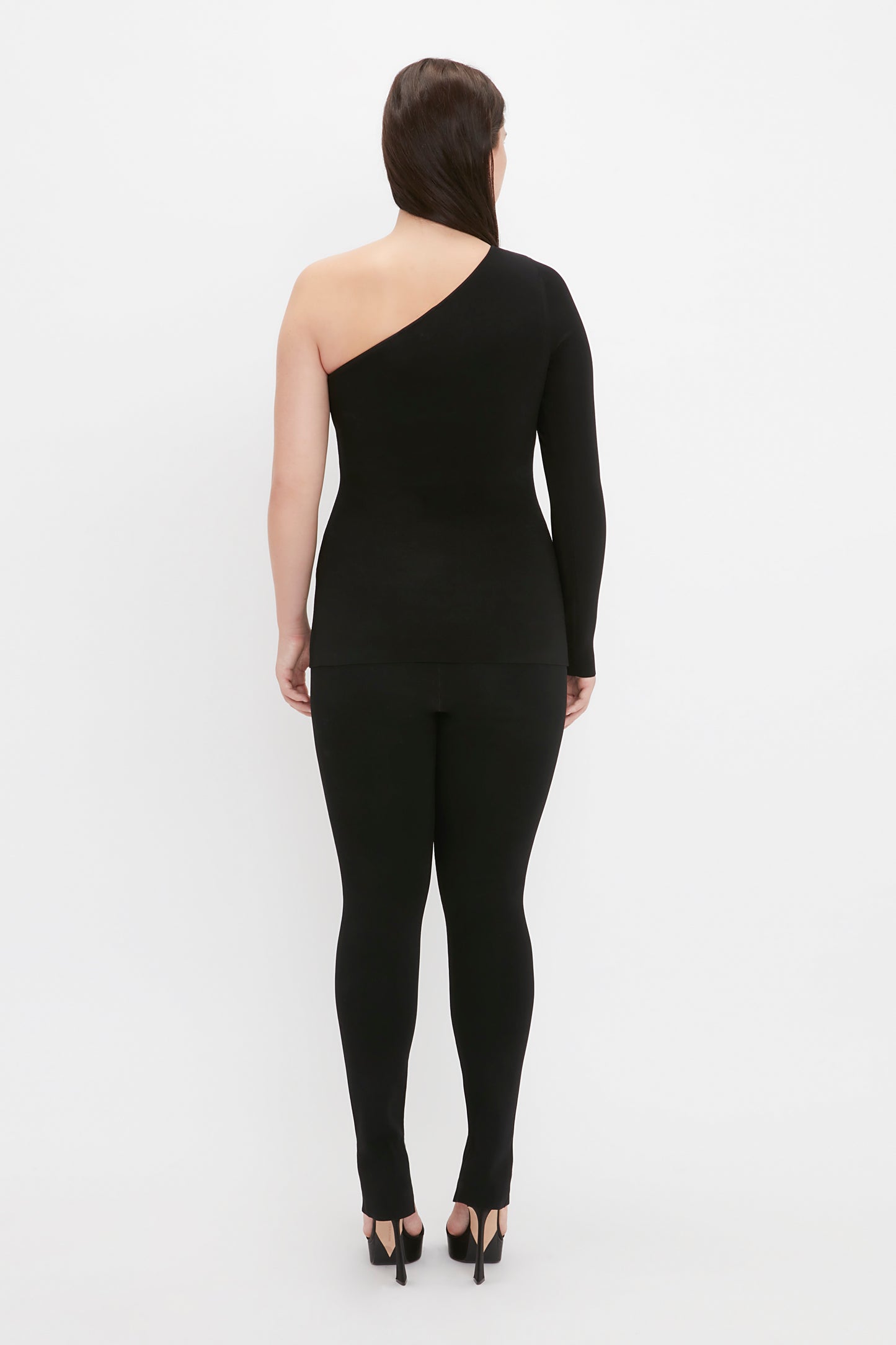 A person stands facing away, wearing the VB Body One Shoulder Top in Black and black fitted pants. They have dark hair and are wearing black high heels. The background is white.