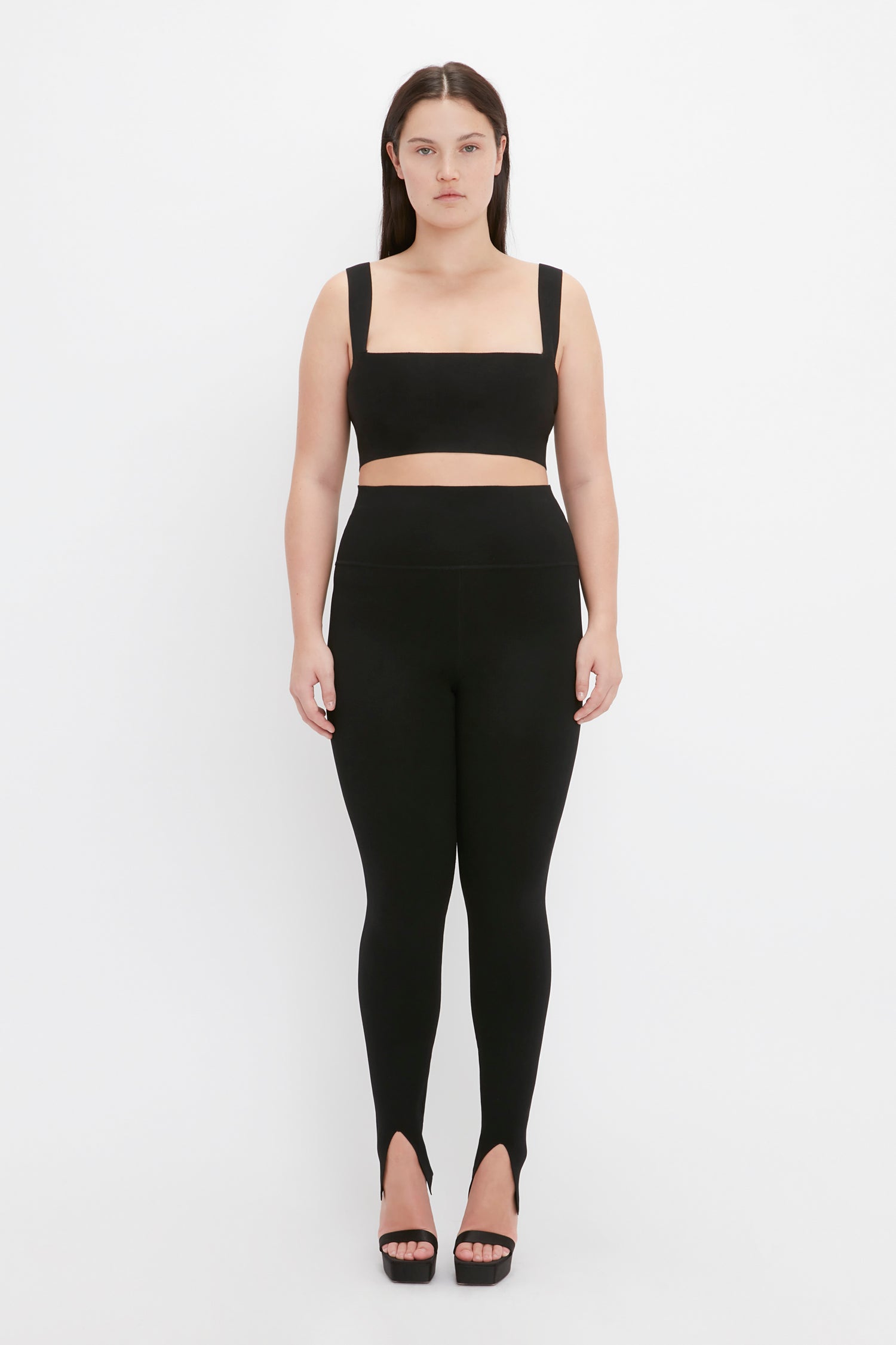 A woman stands against a plain background wearing a black, sleeveless crop top and the VB Body Split Front Leggings in Black by Victoria Beckham with stiletto heels.