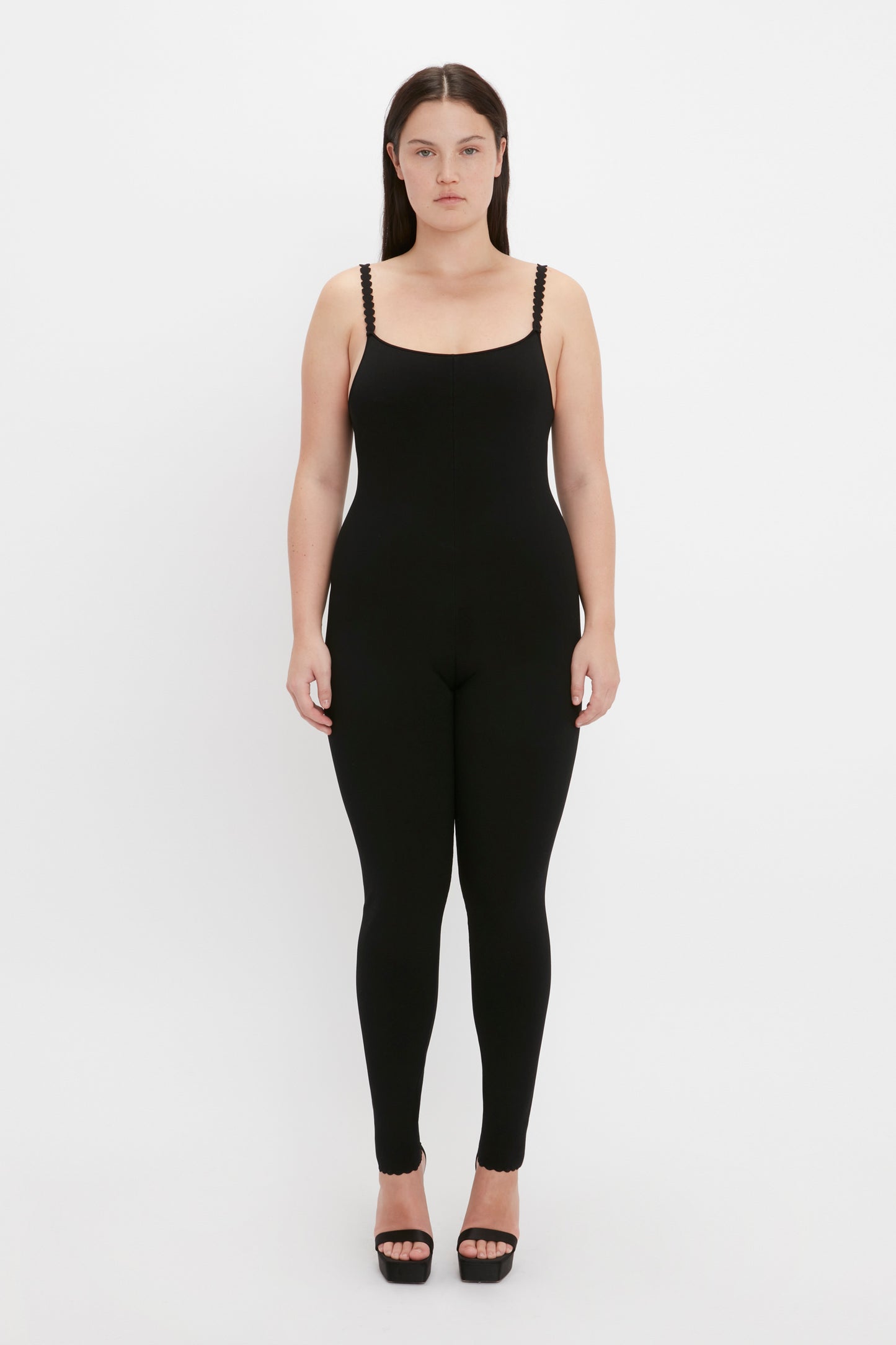 A person stands against a plain white background, wearing the VB Body Catsuit in Black from Victoria Beckham and black high-heeled sandals.