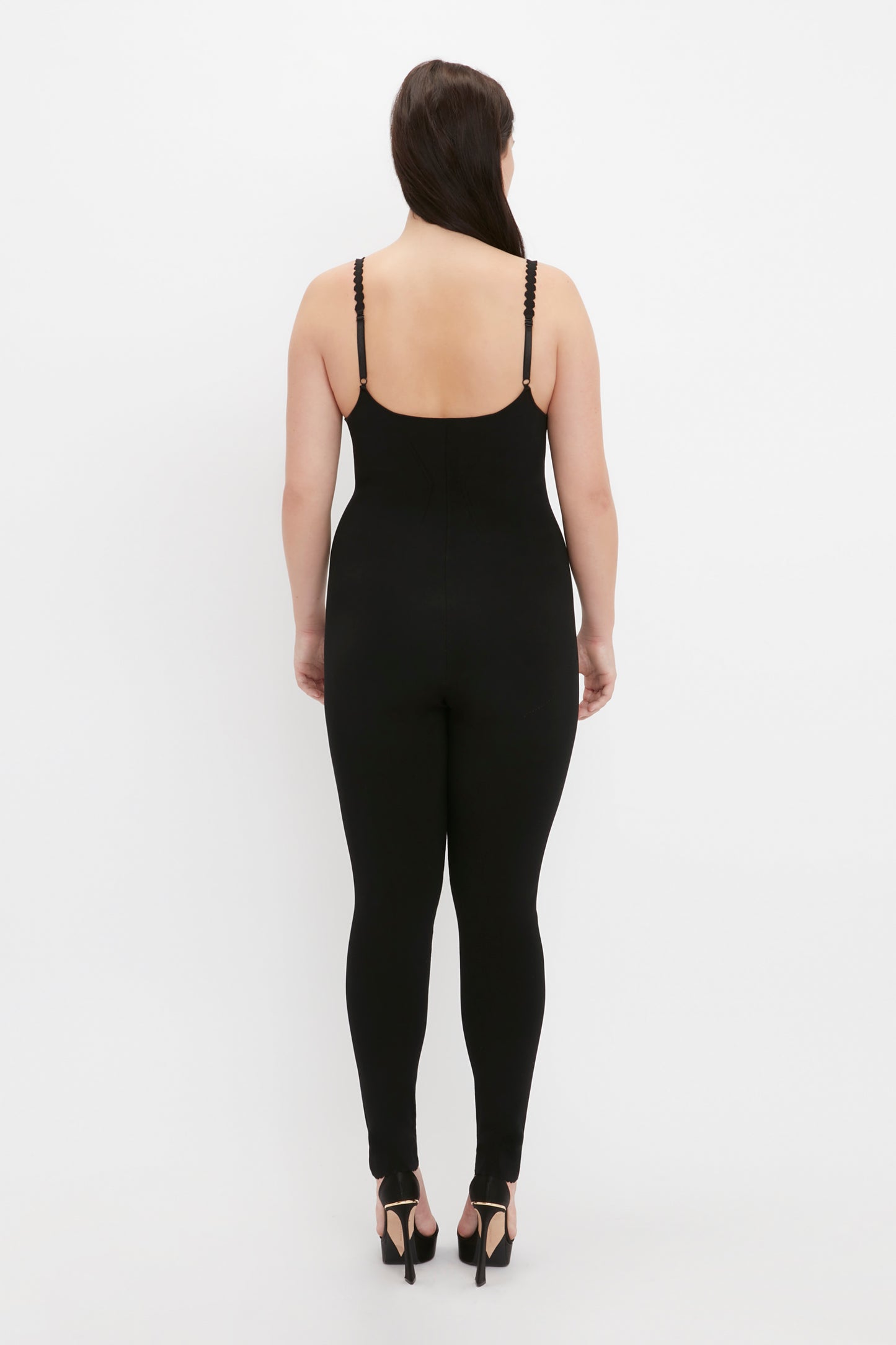 A person in the VB Body Catsuit in Black by Victoria Beckham and heels stands facing away from the camera against a plain white background.