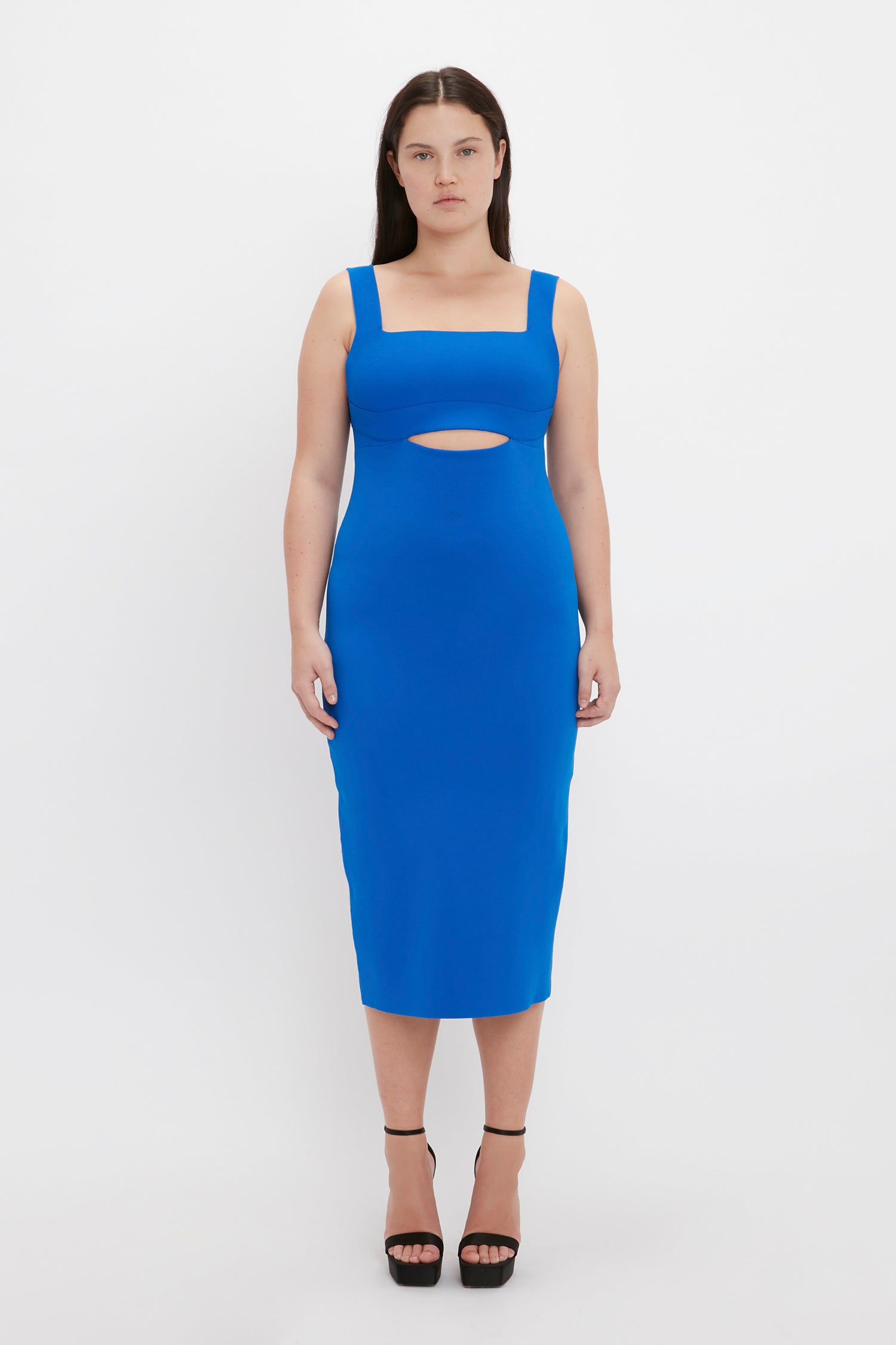 A woman stands facing forward wearing the VB Body Cut Out Midi Dress in Cobalt by Victoria Beckham, paired with black high-heeled sandals.