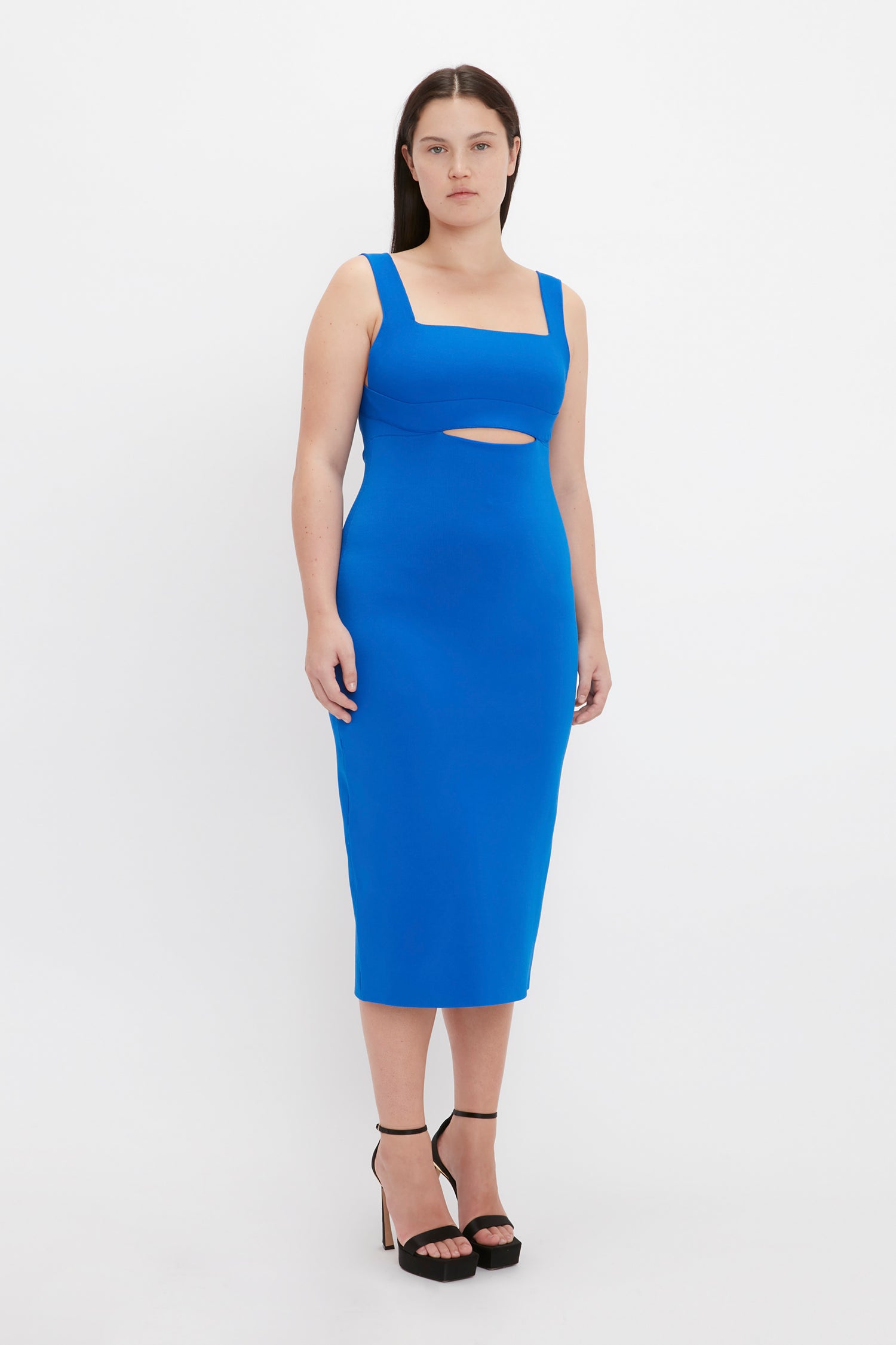 Person standing against a white background wearing the Victoria Beckham VB Body Cut Out Midi Dress in Cobalt and black high heels.