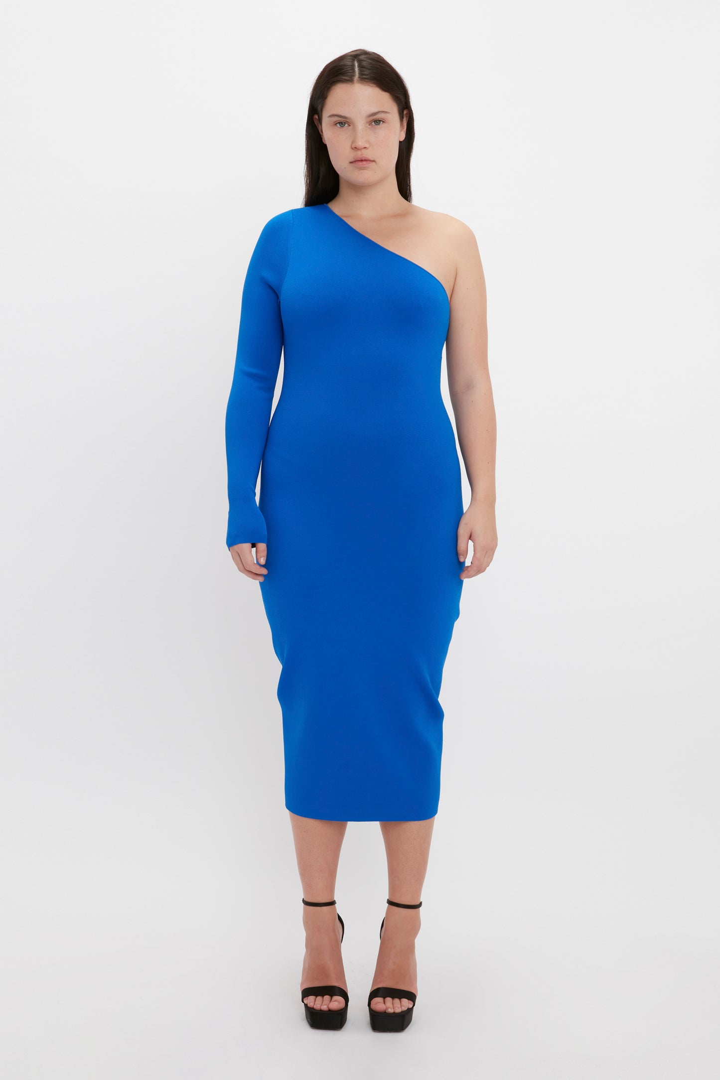 A woman stands against a white background, showcasing the form-fitting VB Body One Shoulder Midi Dress in Cobalt by Victoria Beckham, paired with black high-heeled shoes.
