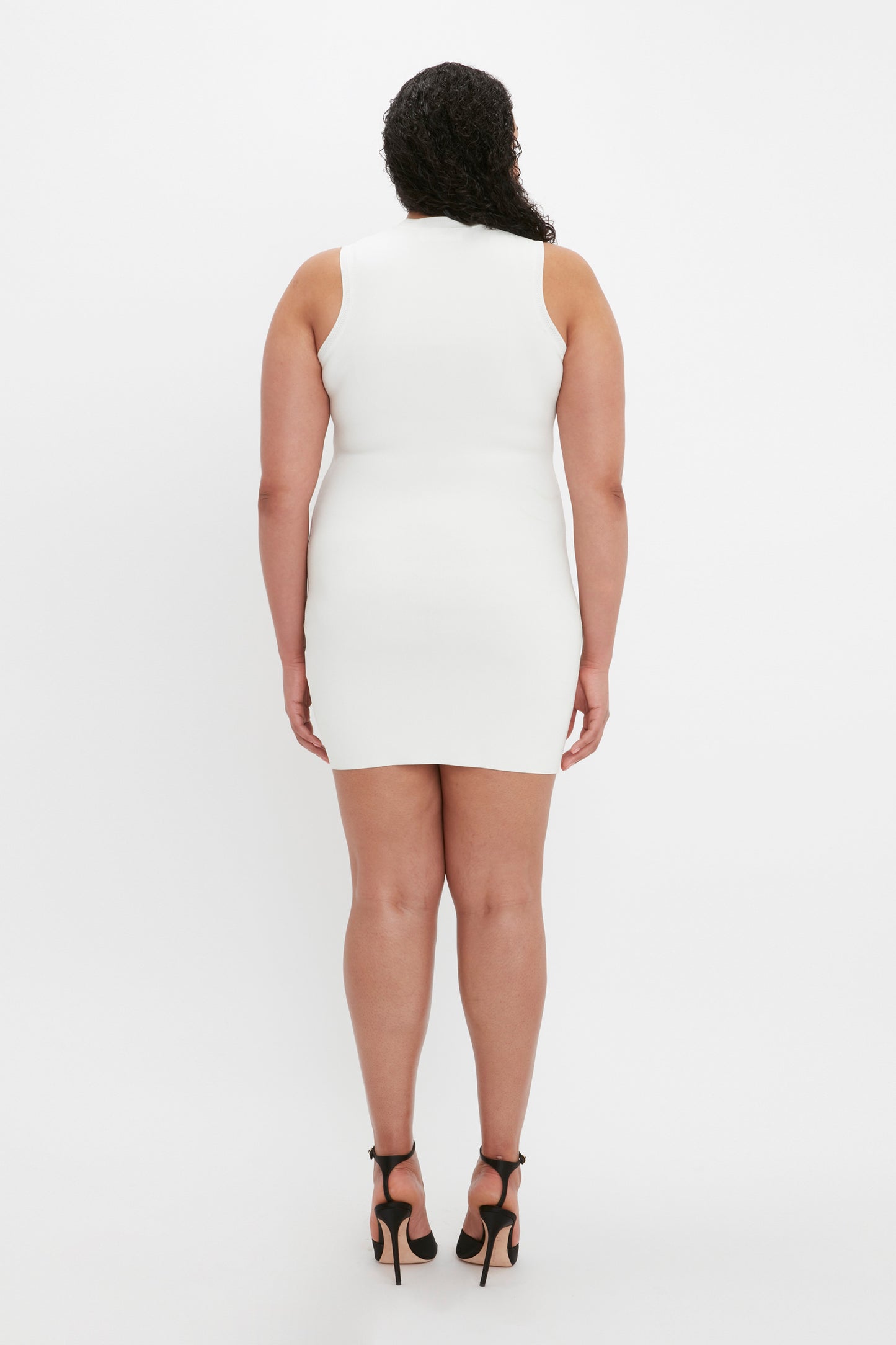 A person with curly hair stands facing away, wearing the VB Body Mini Dress in White from Victoria Beckham and black high heels against a plain white background.