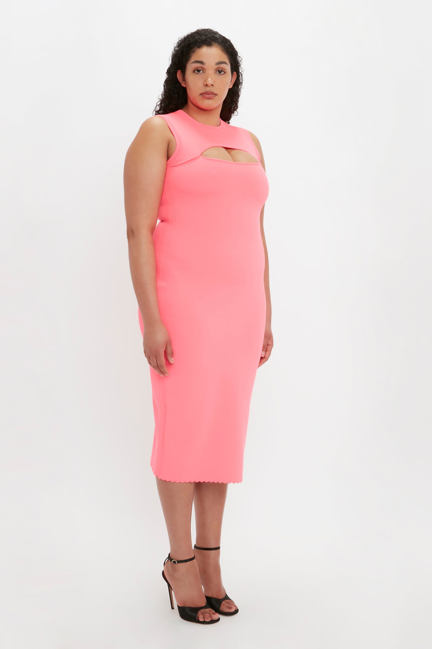 A woman with curly hair stands wearing the Victoria Beckham VB Body Slash Front Midi Dress in Pink and black high-heeled sandals.