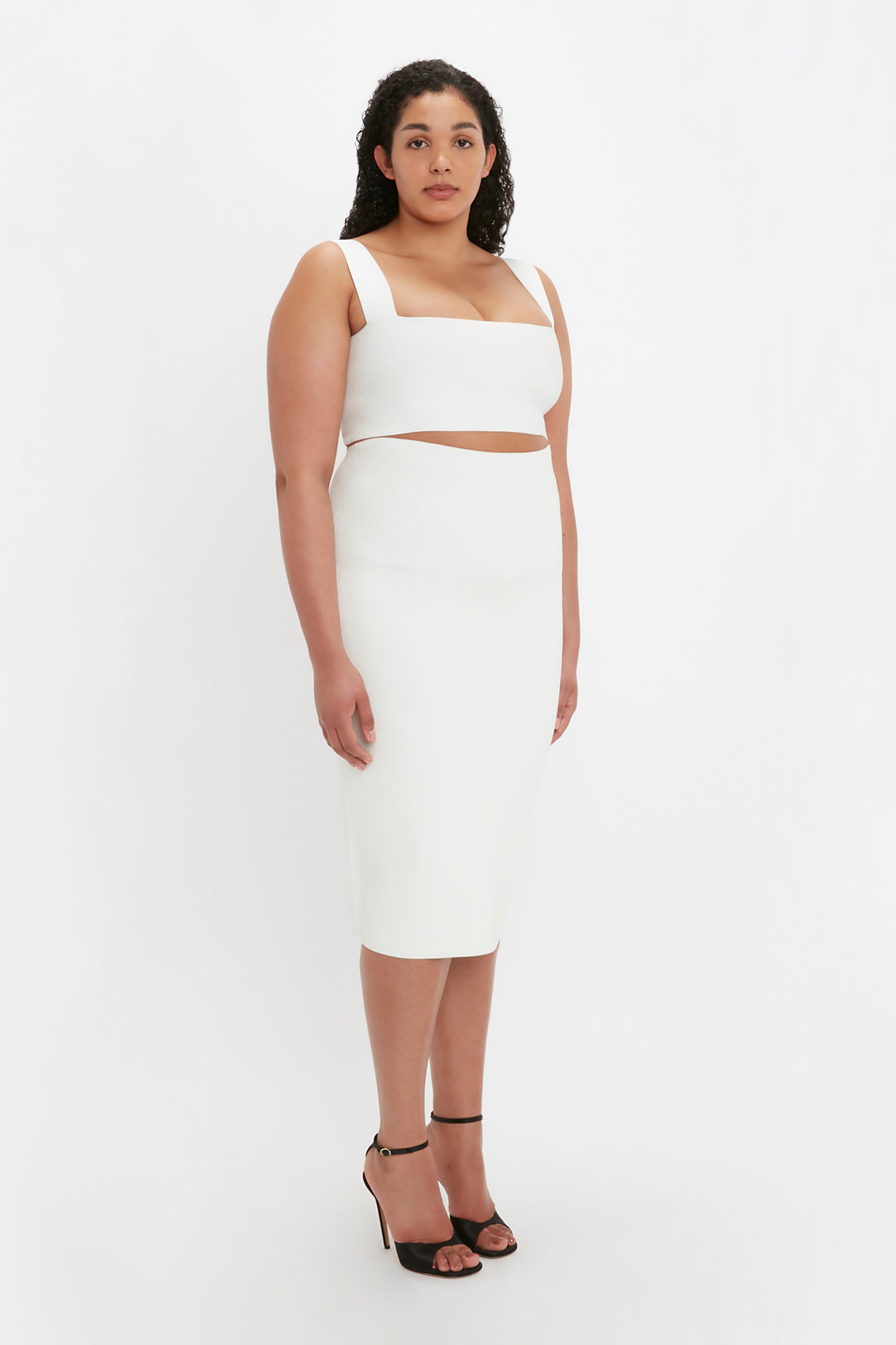 Person standing against a white background, wearing the VB Body Strap Bandeau Top in white by Victoria Beckham, along with a white sleeveless dress featuring a midriff cutout and black high-heeled sandals.