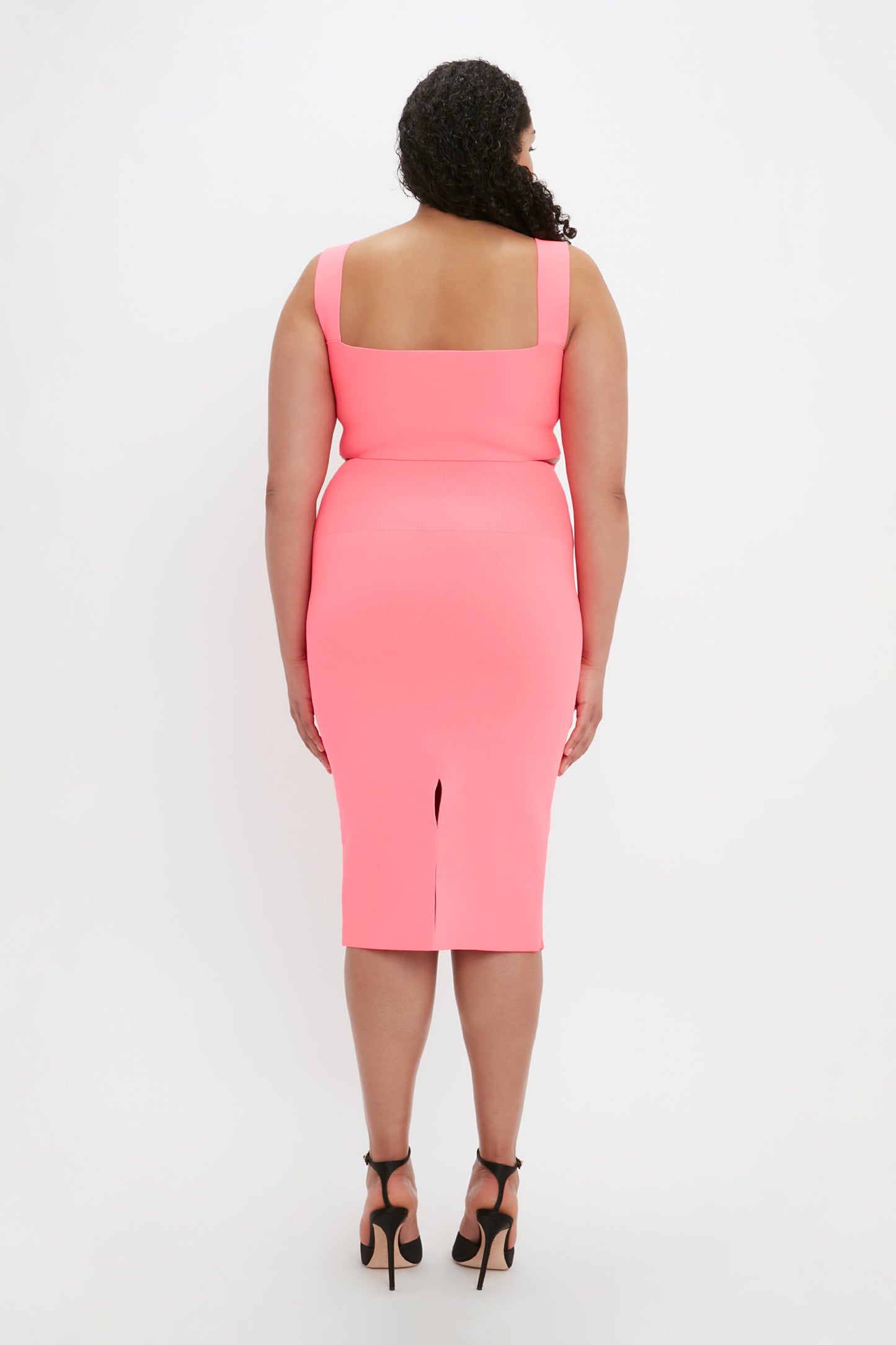 A person stands facing away from the camera, wearing a pink VB Body Strap Bandeau Top by Victoria Beckham and black high-heeled shoes.