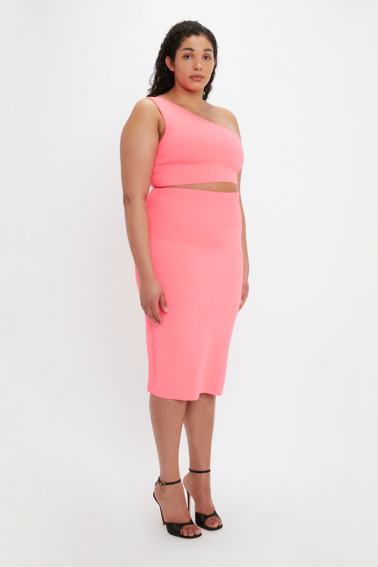 A person stands against a plain background, wearing the VB Body One Shoulder Crop Top in Pink by Victoria Beckham, paired with a matching asymmetrical bottom and black high-heeled sandals.