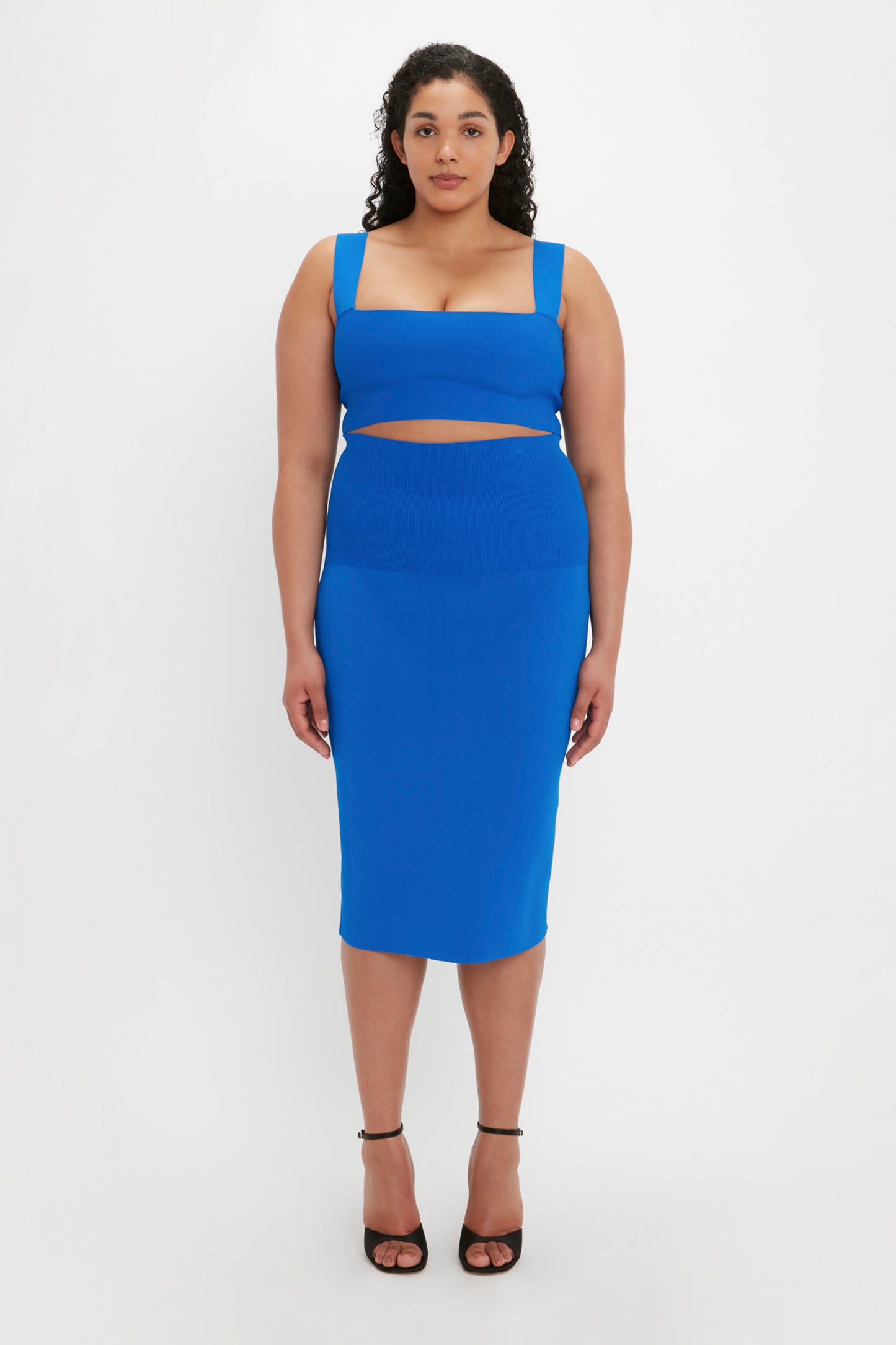 A woman wearing a Victoria Beckham VB Body Strap Bandeau Top in Cobalt and black high heels stands against a plain white background.