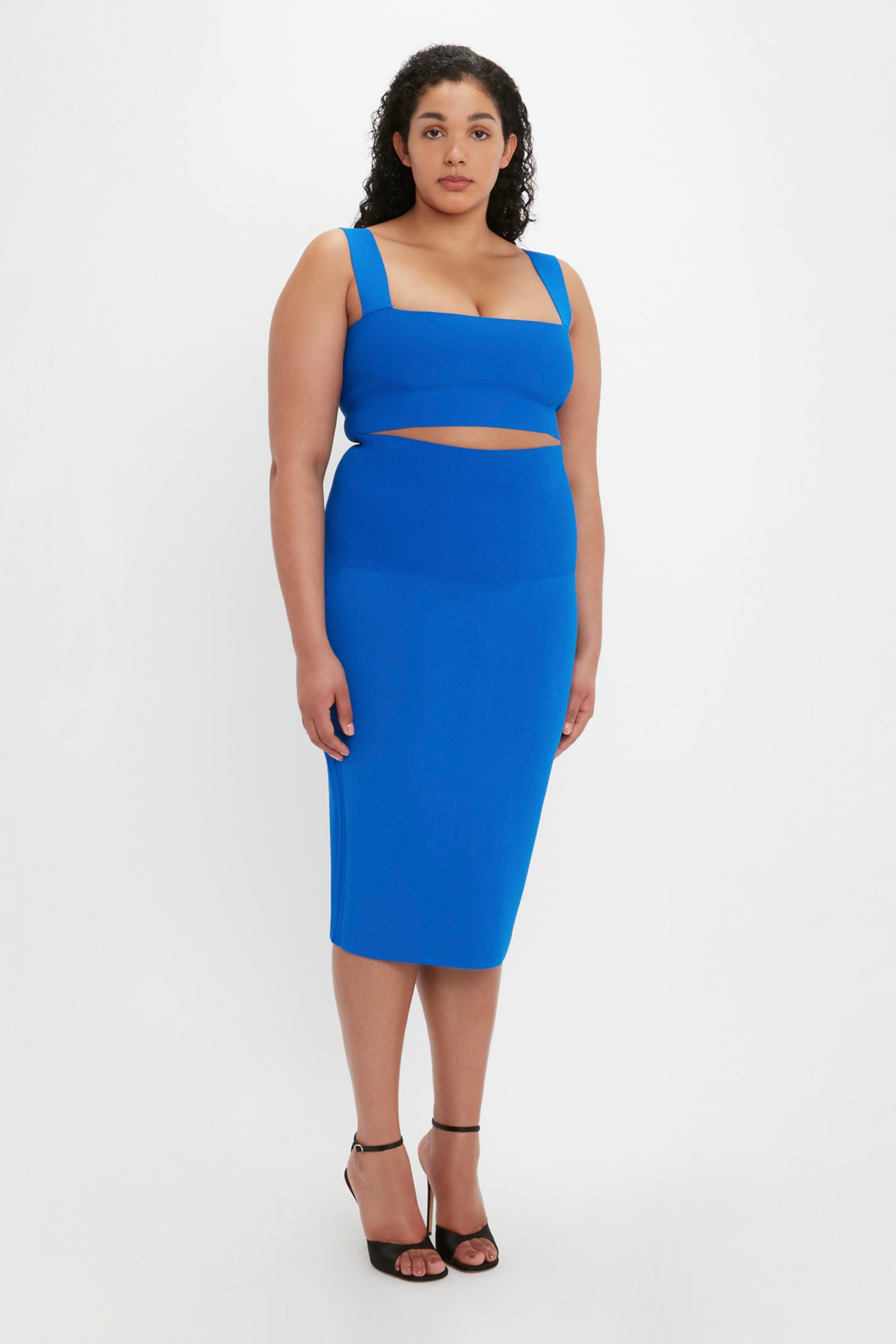 A woman with curly hair stands against a white background, wearing a blue sleeveless crop top, the Victoria Beckham VB Body Fitted Midi Skirt in cobalt, and black heeled sandals.