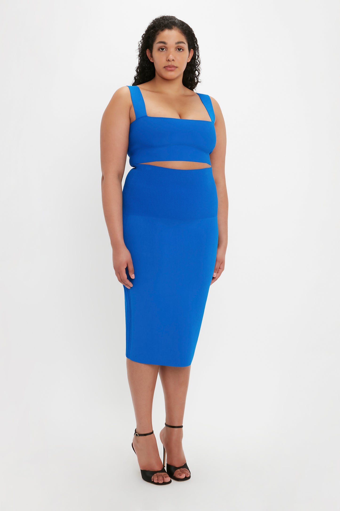 A person with curly hair stands against a plain white background, wearing the Victoria Beckham VB Body Strap Bandeau Top in Cobalt paired with matching blue bottoms and black heels.