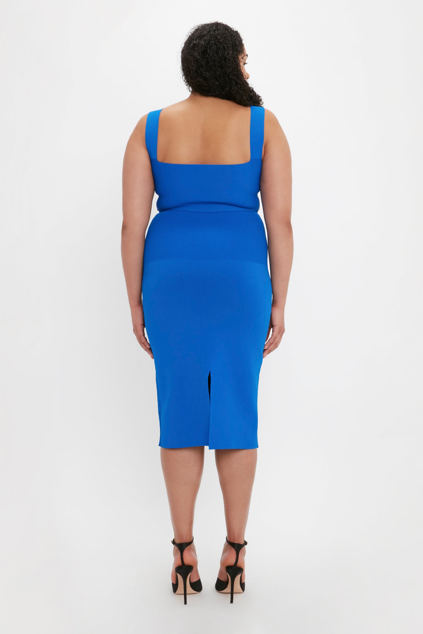 A woman with curly hair is standing with her back facing the camera, wearing a blue Victoria Beckham VB Body Fitted Midi Skirt in Cobalt and black high-heeled shoes.
