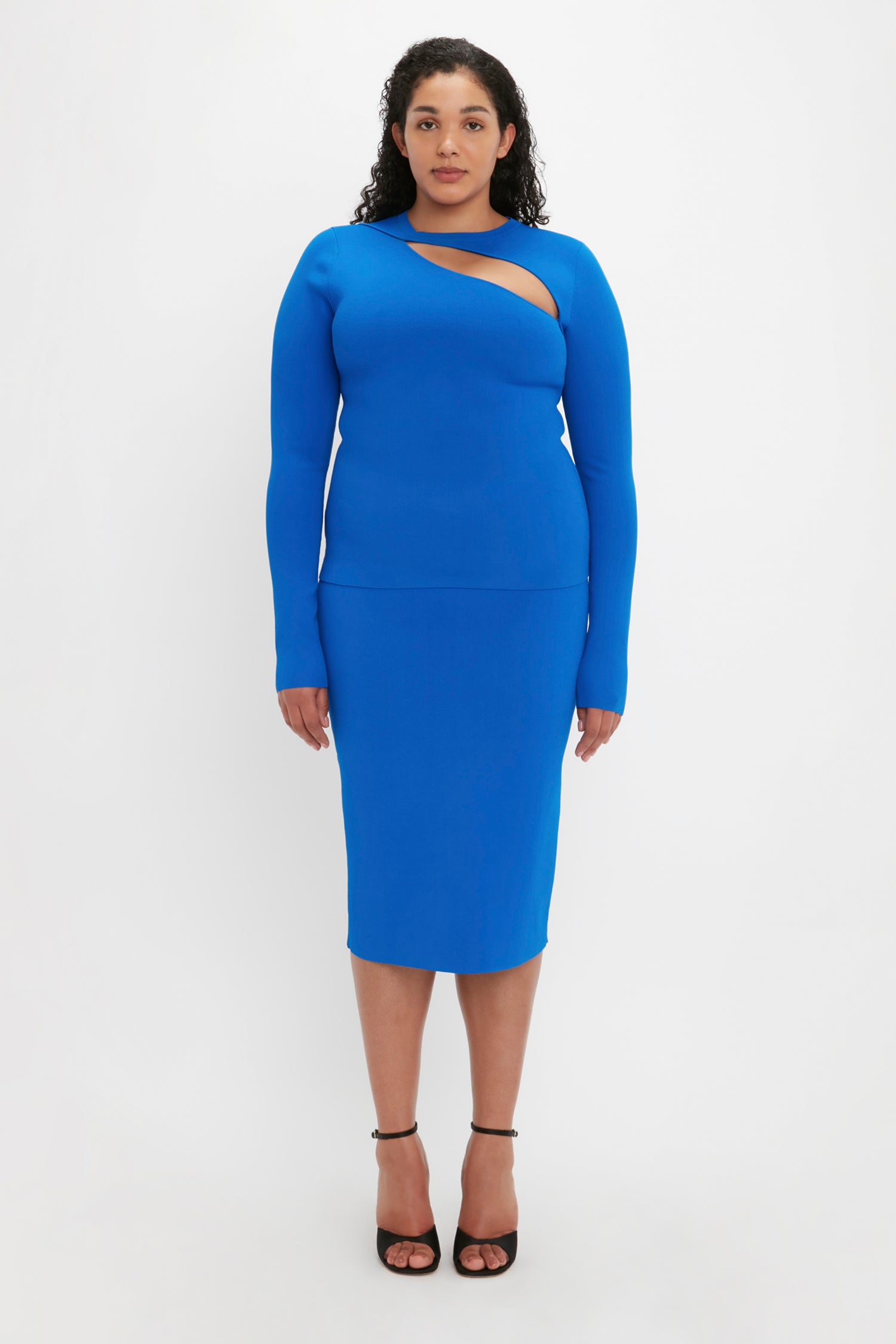 A person stands against a white background wearing the Victoria Beckham VB Body Front Slash Crew Neck Top in Cobalt and black high-heeled sandals.
