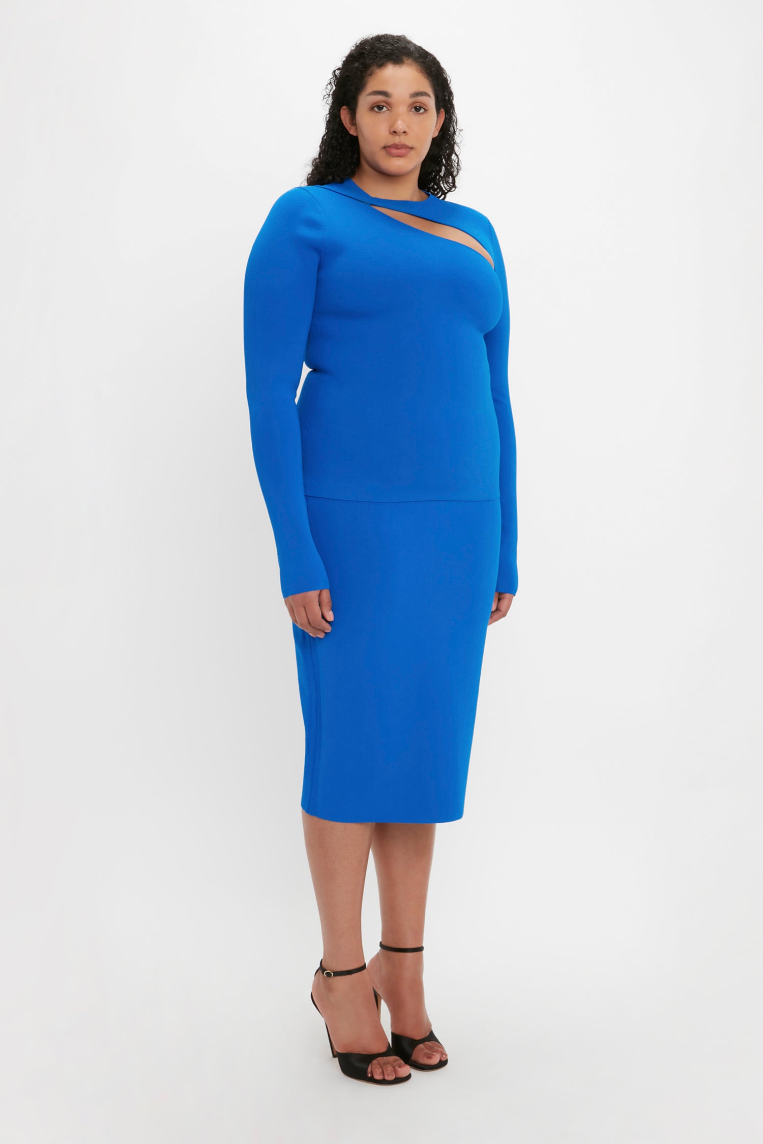 A woman stands in a Victoria Beckham VB Body Front Slash Crew Neck Top in Cobalt, paired with black heels, posing against a plain white background.